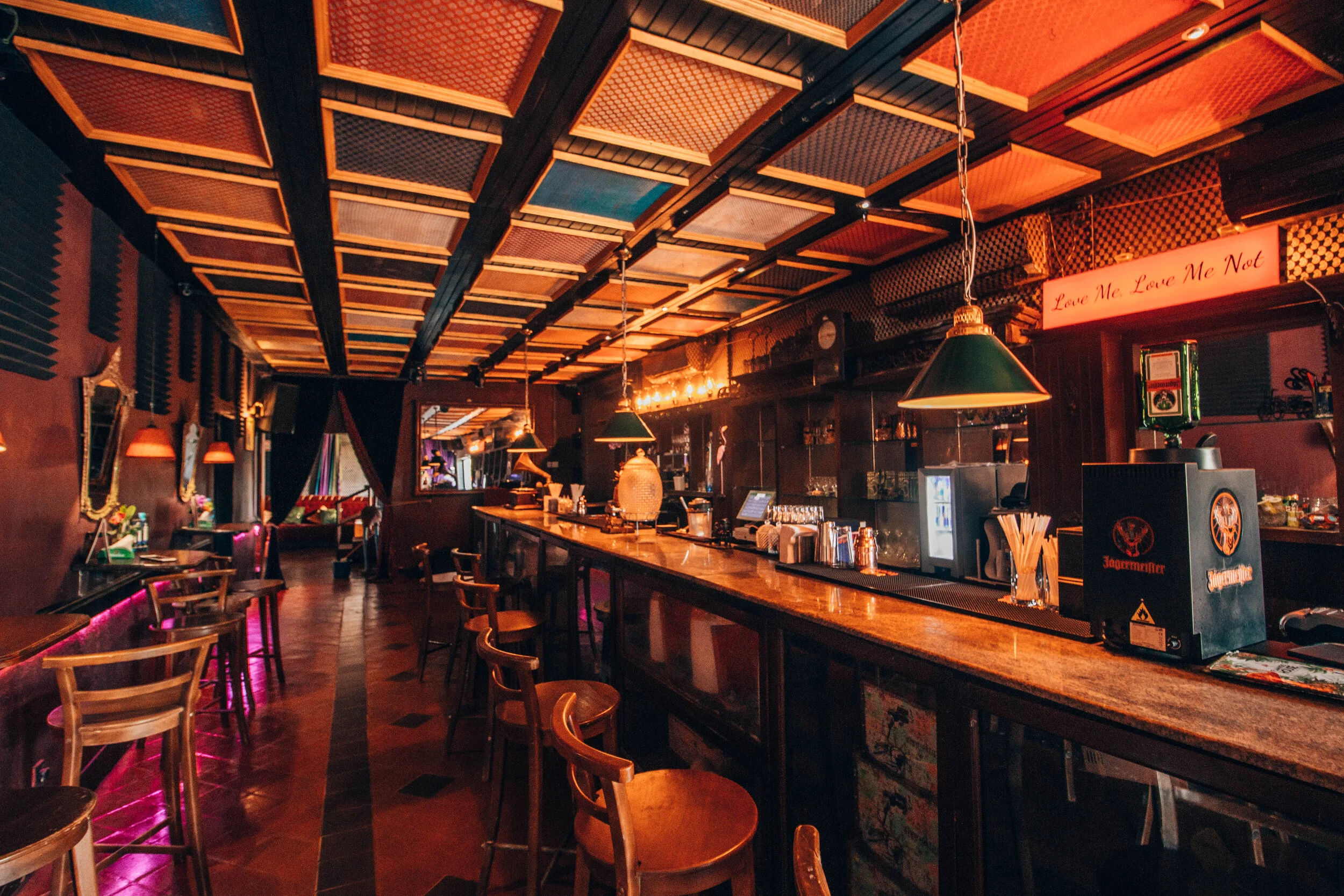 Interior of a dimly lit bar with a long wooden counter, wooden chairs, sipping glasses, green hanging lamps, and decorative walls with black acoustic panels and mirrors.