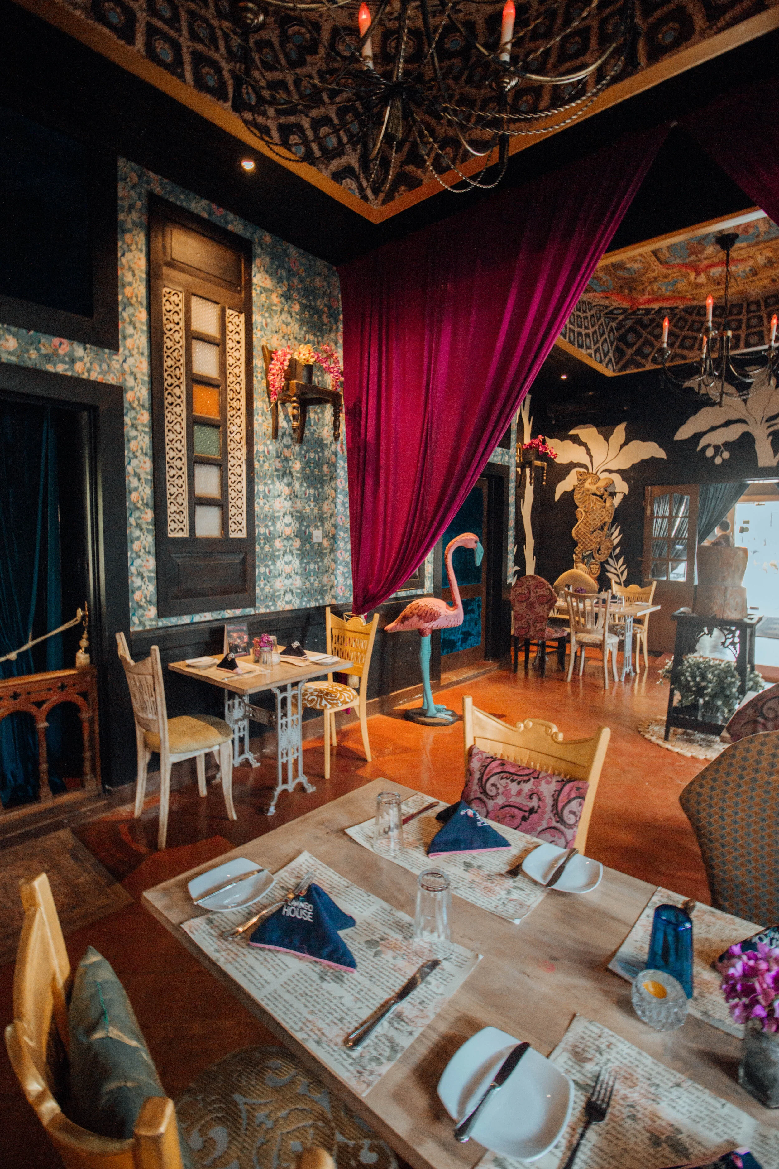 Decorative restaurant interior with floral wallpaper, pink flamingo statue, red velvet curtain, chandelier, and eclectic furniture.