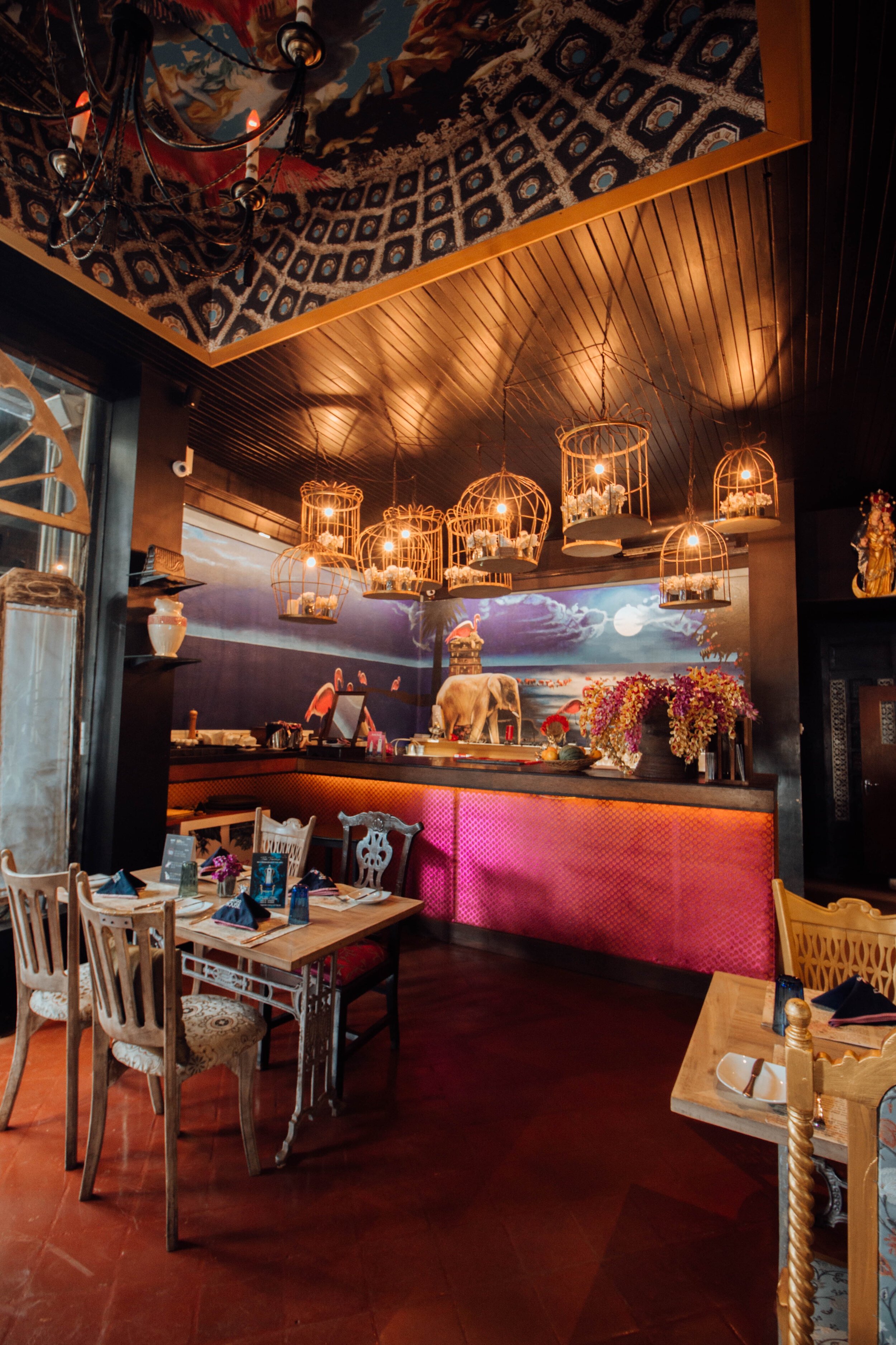 Interior of a restaurant or cafe with a tropical-themed painted mural of an elephant, palm trees, and a sunset or sunrise in the background. The ceiling has decorative lighting fixtures resembling birdcages with light bulbs inside. The restaurant has