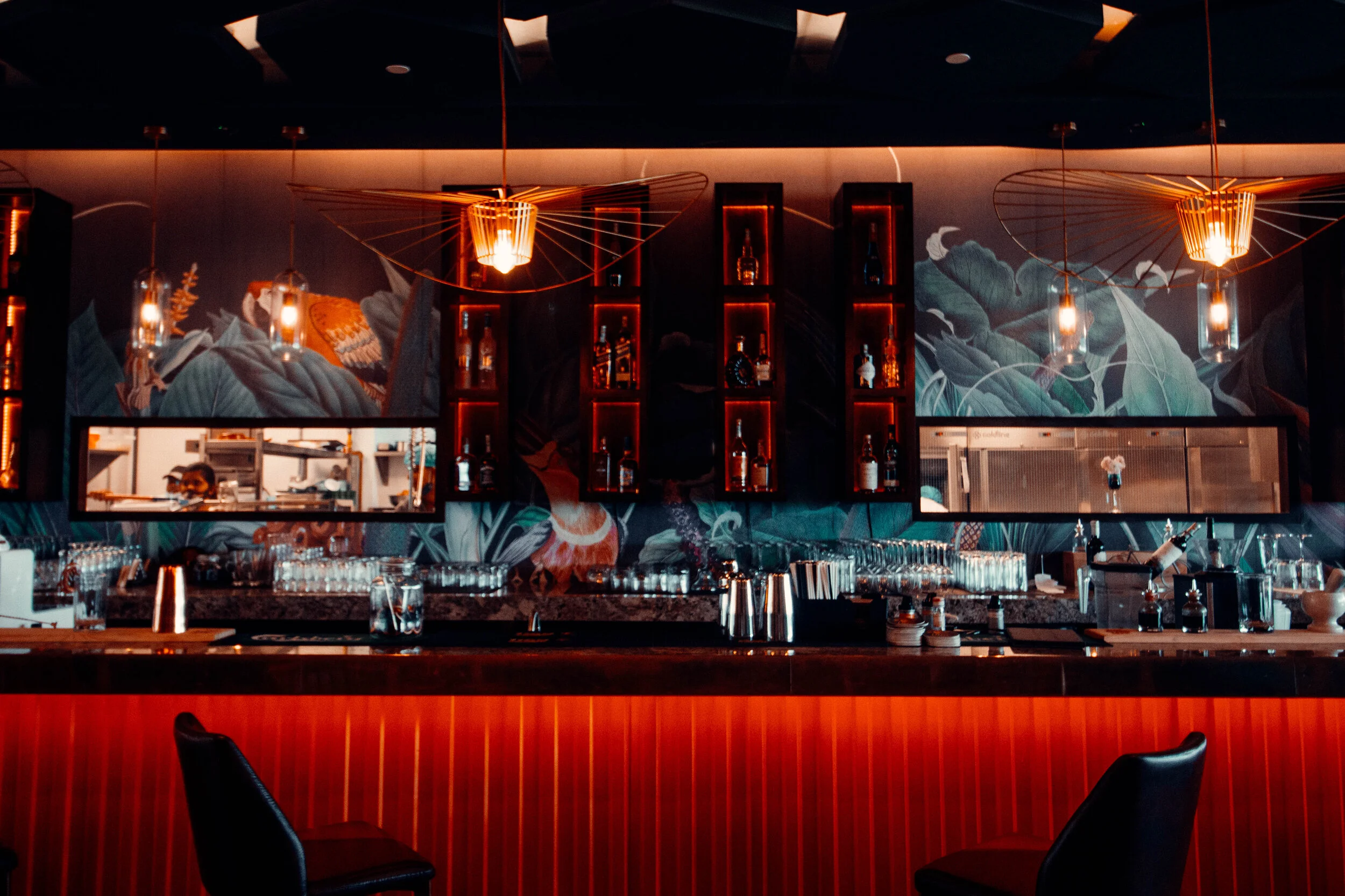 A dimly lit bar with modern decor, featuring illuminated bottles on wall shelves, hanging pendant lights, and a counter with glasses and bar tools.