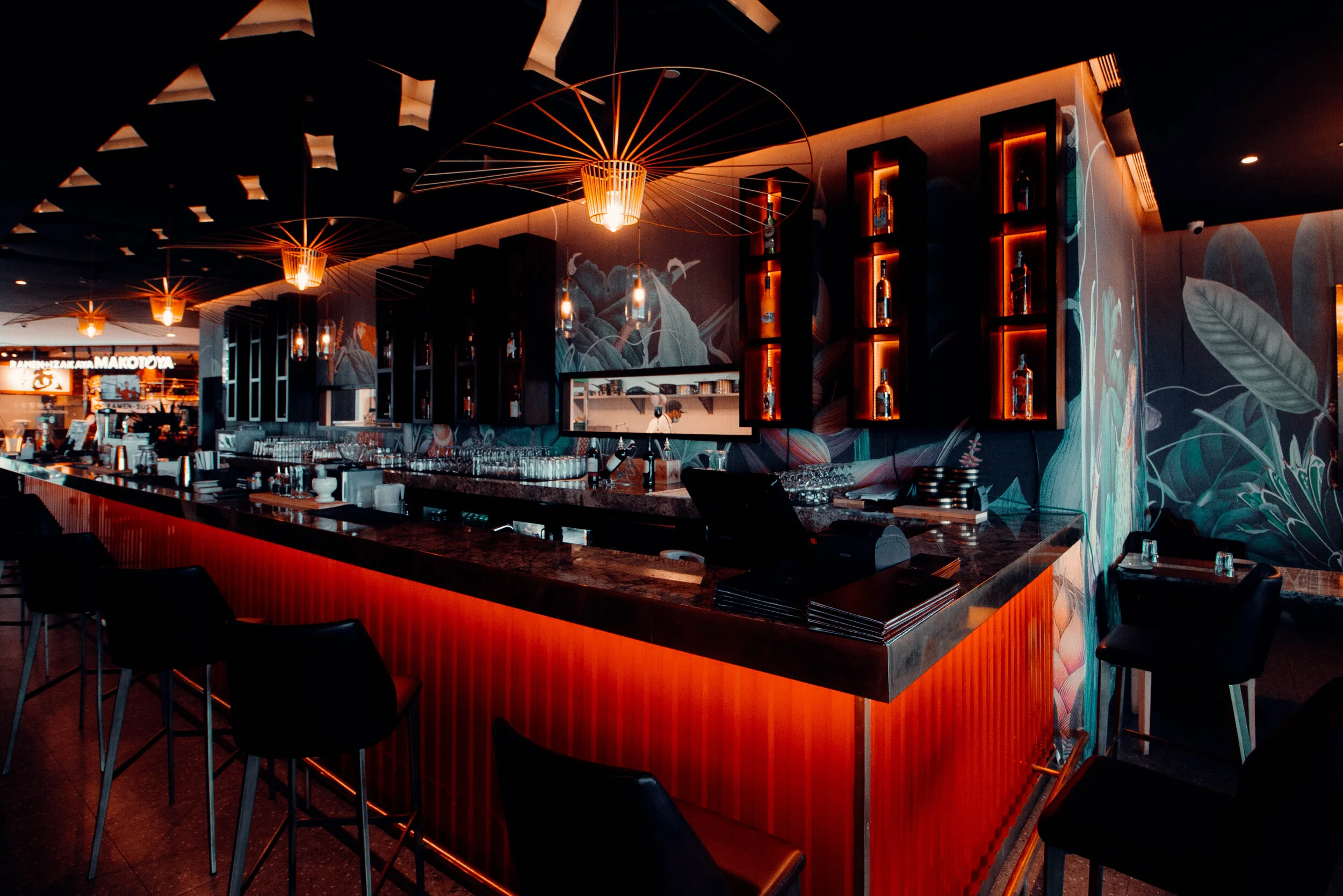 Interior of a stylish bar with dim lighting, illuminated by hanging pendant lights, featuring a marble countertop, black chairs, decorative shelves with bottles, and a vibrant mural with tropical leaves and animals.
