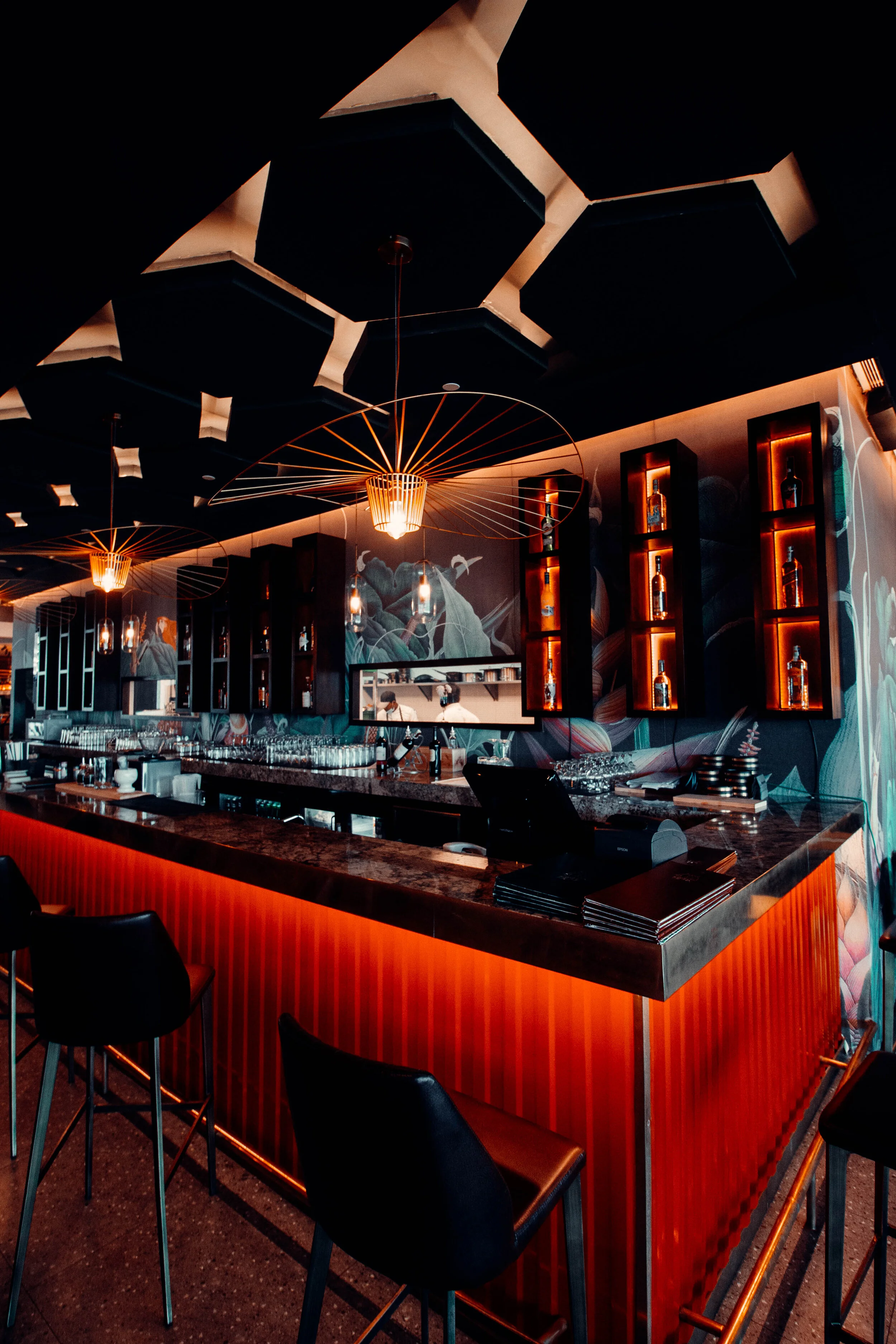 Dimly-lit modern bar with orange and black decor, featuring hanging decorative lighting fixtures, shelves with bottles, and bar stools.