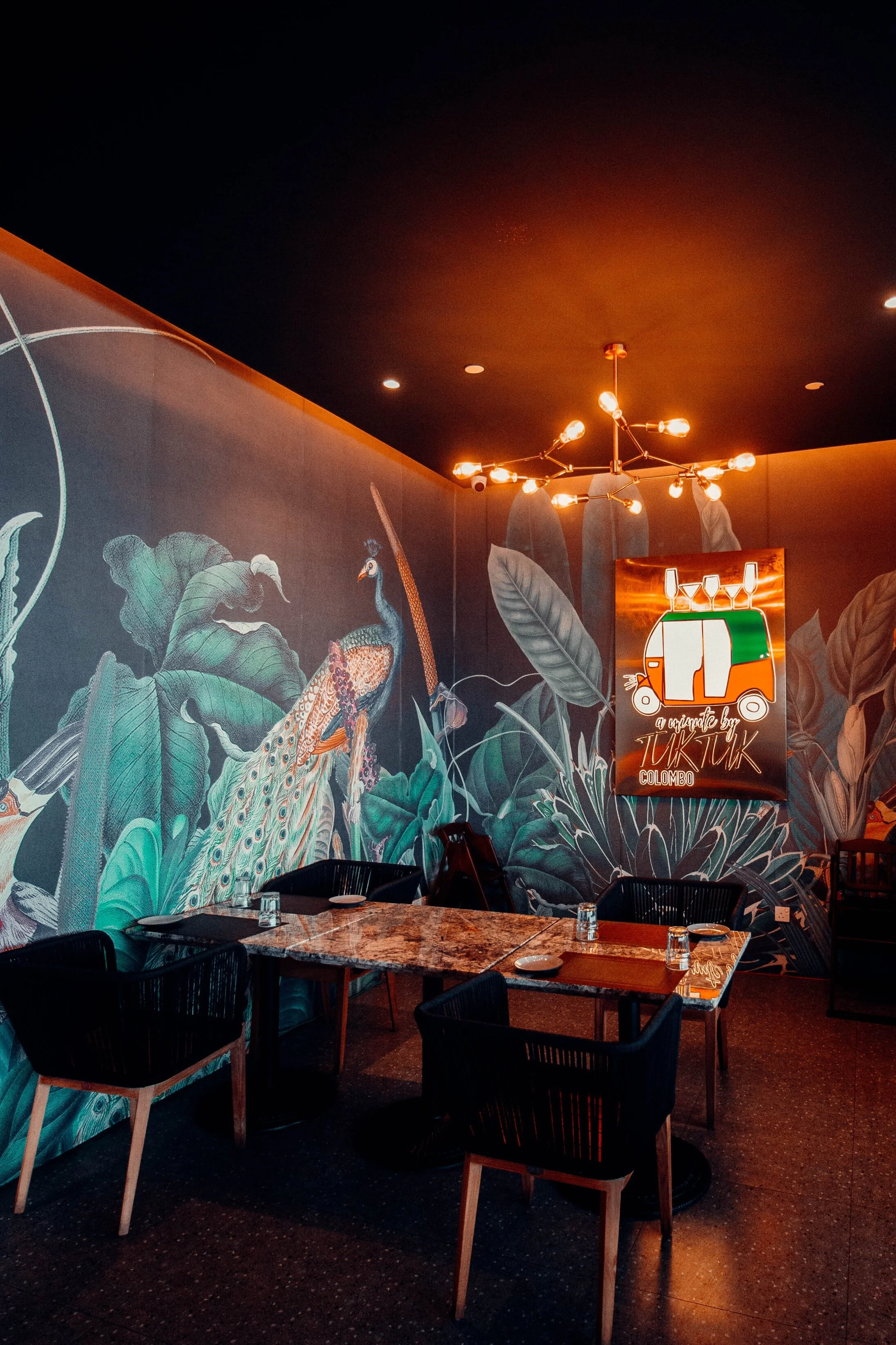 Interior of a restaurant with dark walls featuring tropical mural art, a modern chandelier, and a colorful sign that reads 'a tribute by TikTik Colombo'.