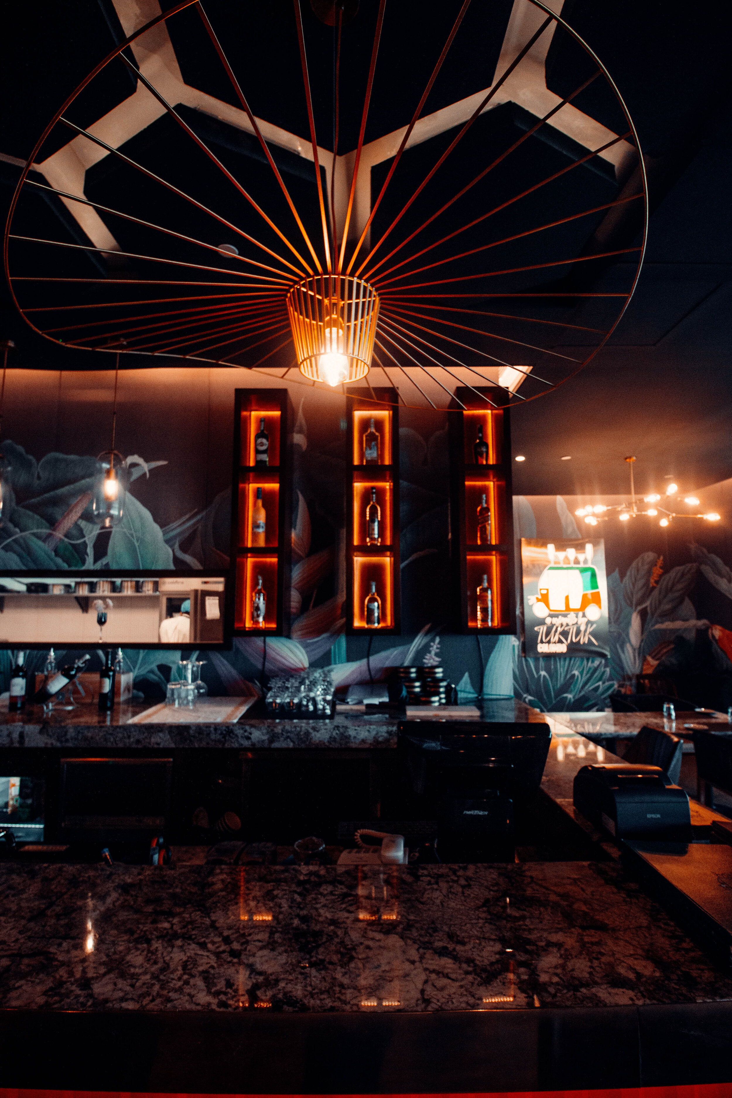 Interior of a bar with a dark aesthetic, featuring hanging pendant lights, a decorative ceiling fixture, illuminated bottles on shelves, and a mural with floral designs on the wall.