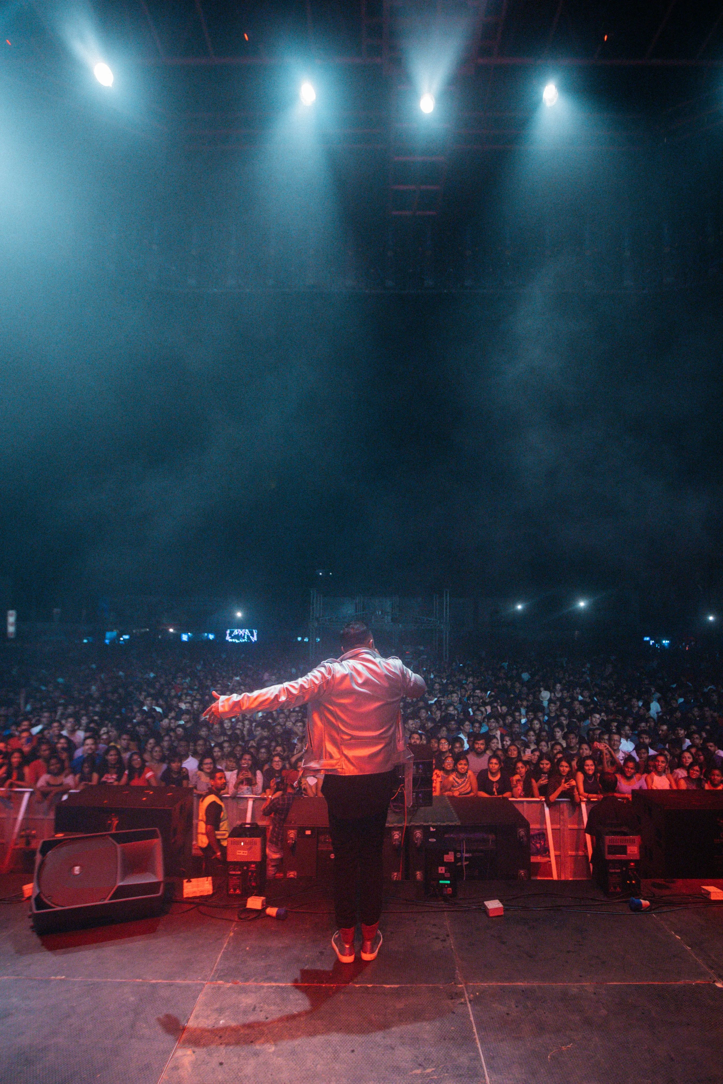 Performer on stage facing a large audience in a concert hall with bright stage lights overhead.
