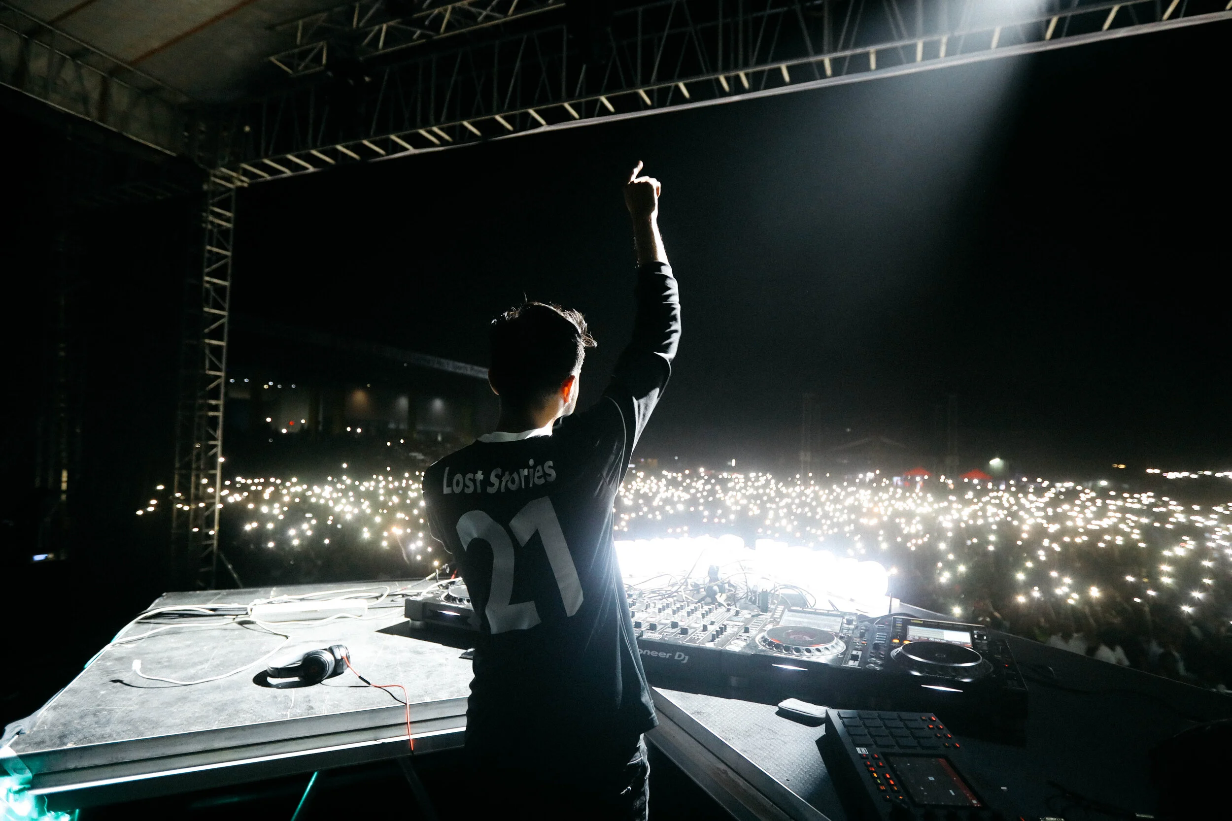 DJ performing on stage with his back to the camera, wearing a black shirt with 'Lost Stories 21' on the back, in front of a large audience at night, filled with lights from phones.