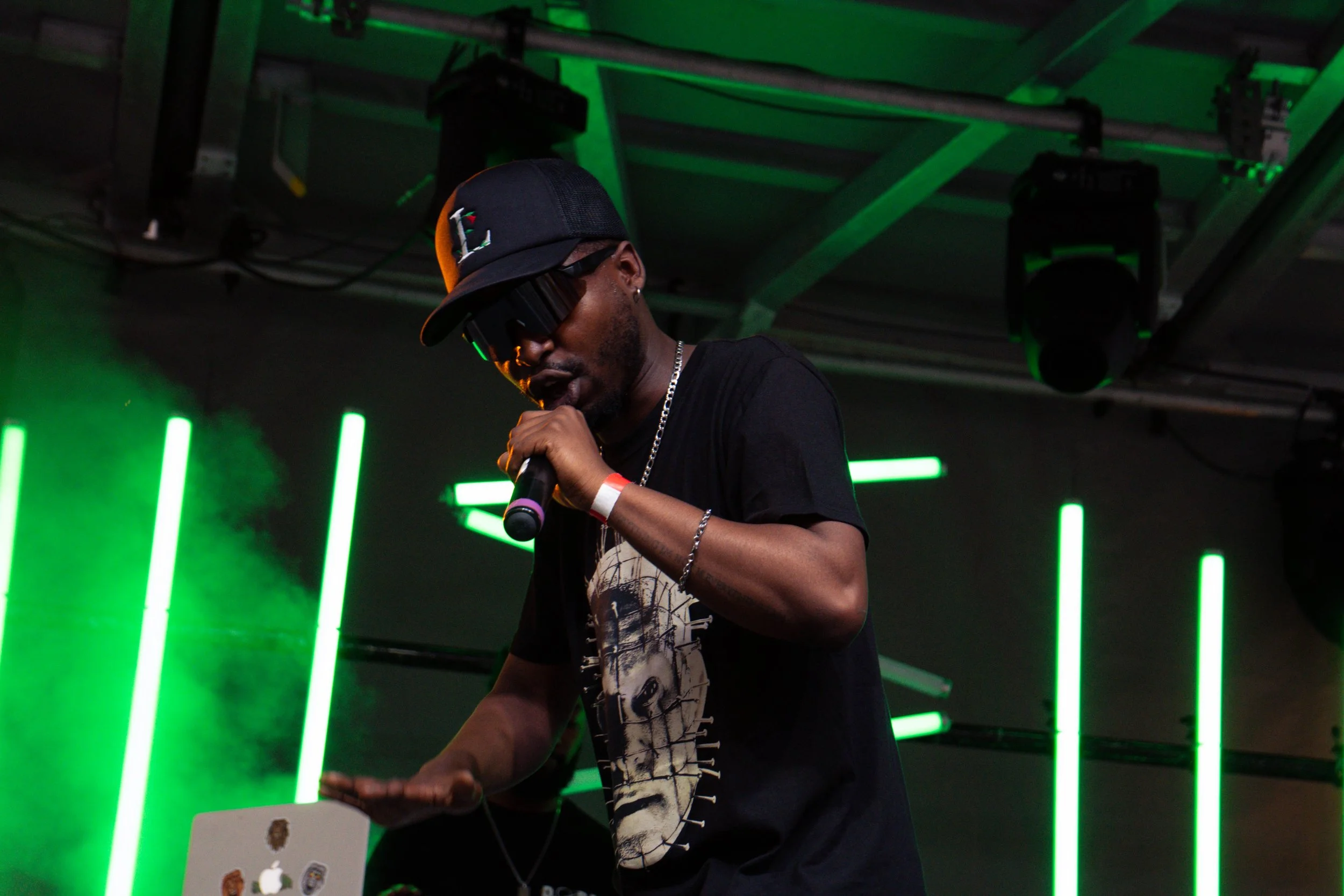 Performing artist wearing a black T-shirt with a graphic design, black baseball cap, and sunglasses, holding a microphone while performing on stage with green neon lights in the background.
