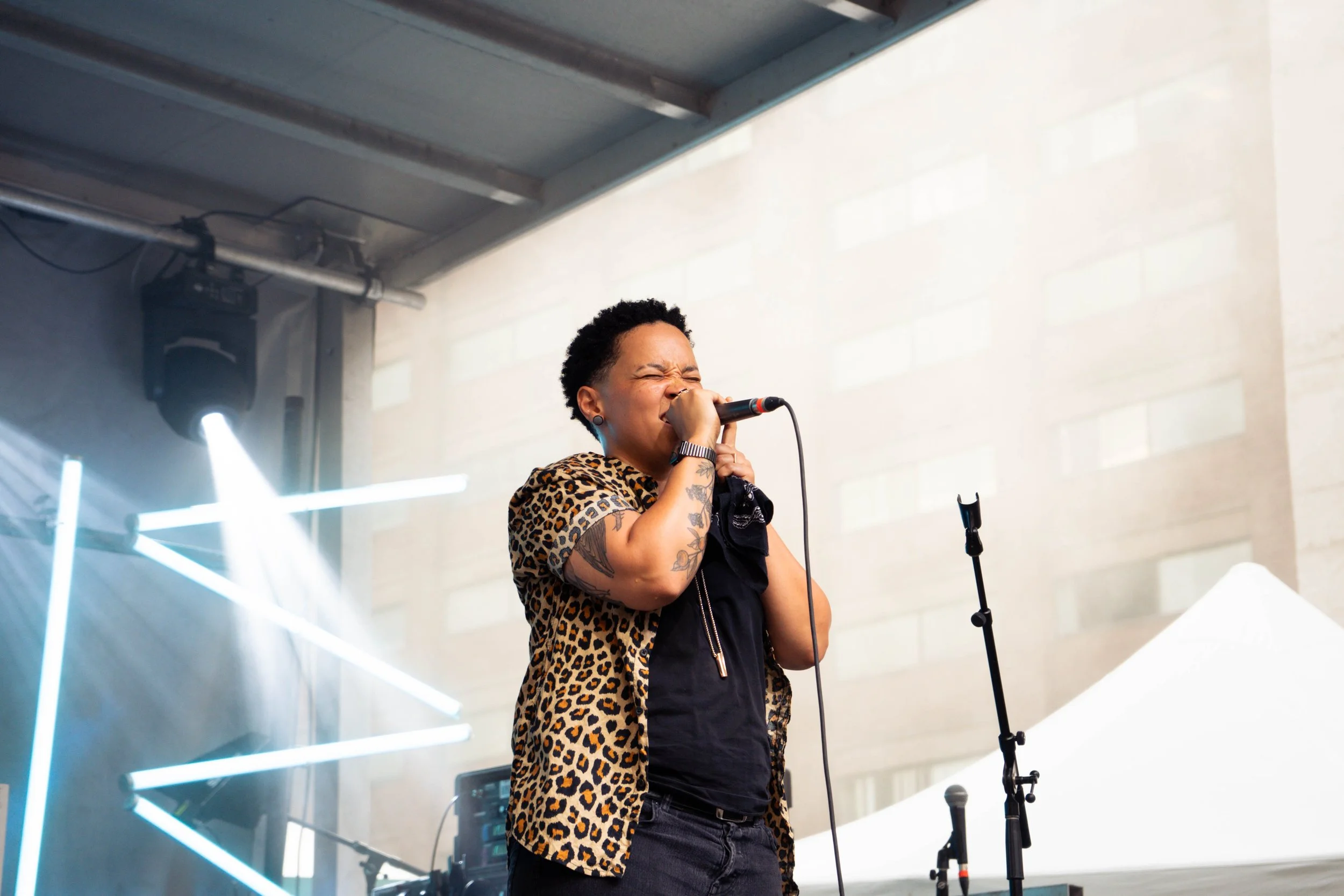 A woman singing into a microphone on stage at an outdoor event, wearing a leopard print shirt and tattoos on her arm, with stage lights and equipment visible in the background.