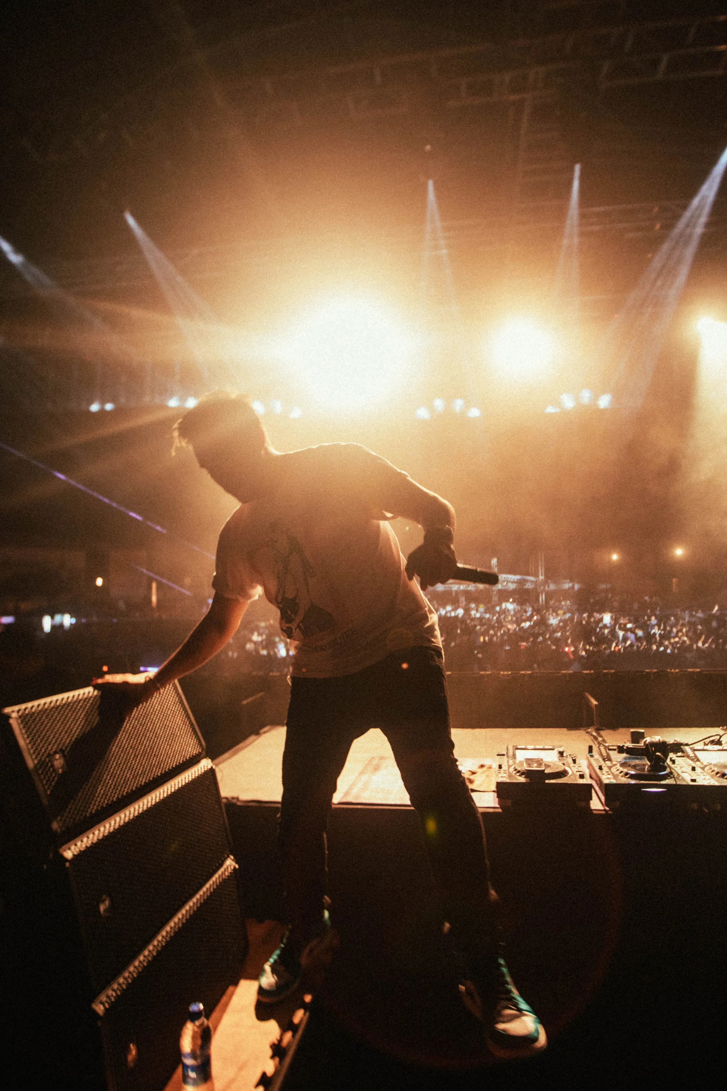 DJ performing on stage with bright lights and large crowd in the background at a concert or event.