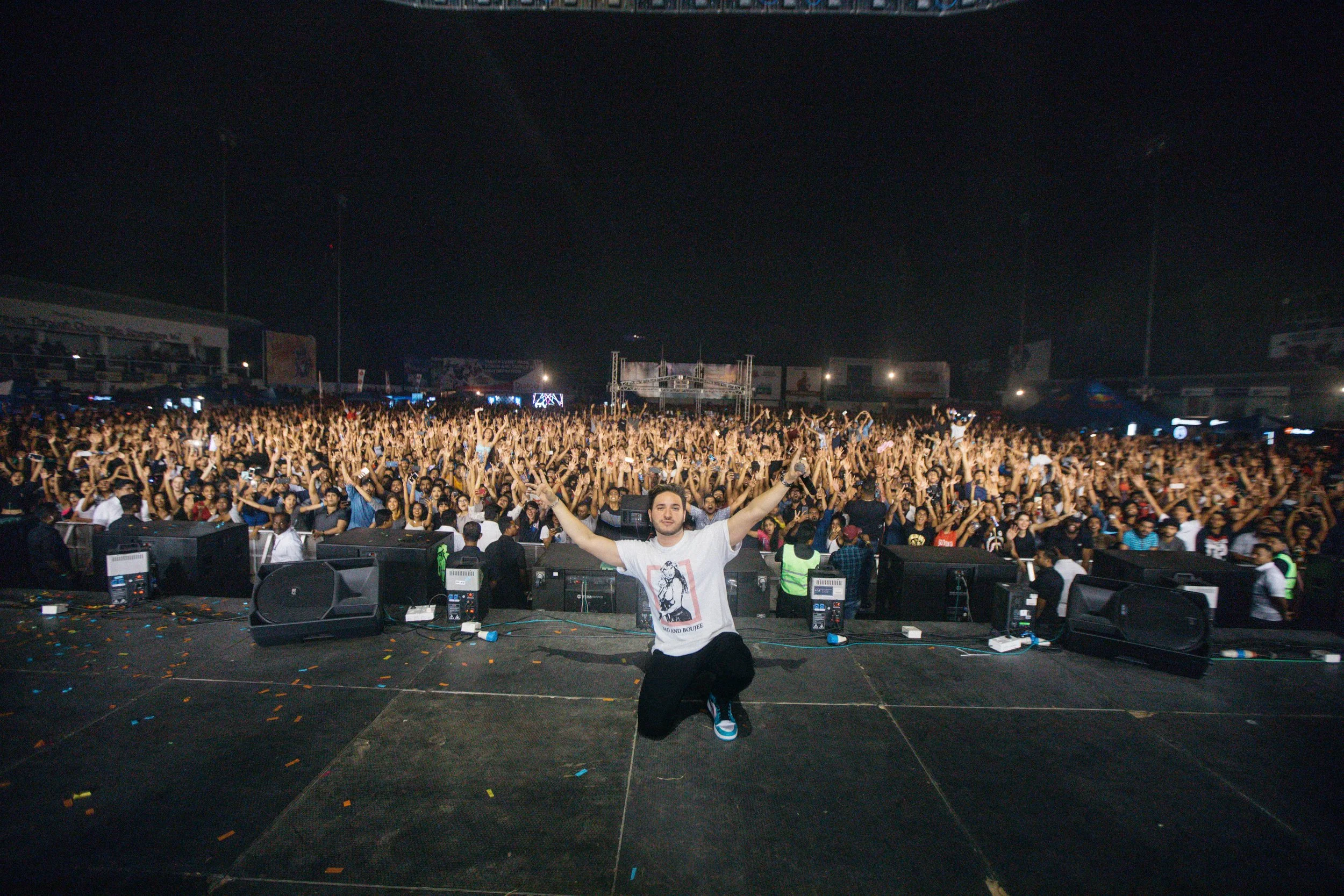 A man poses with arms outstretched on stage in front of a large crowd at a concert or festival during the night, with the audience raising their hands.