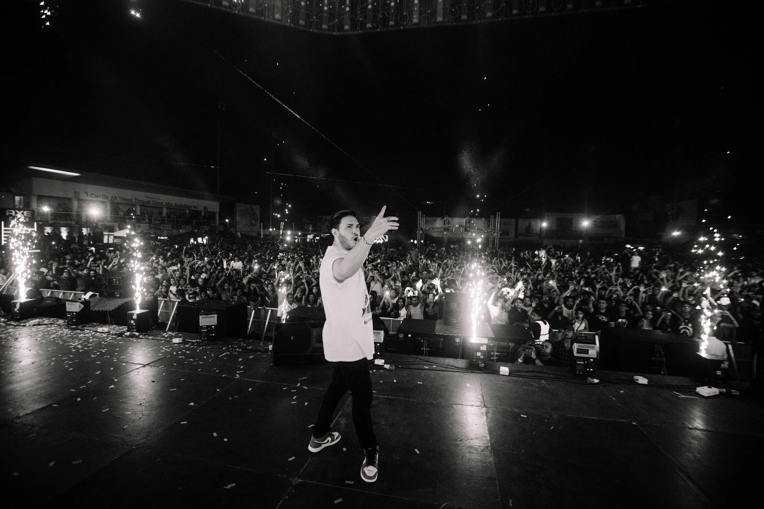 A performer on stage singing in front of a large crowd at a nighttime concert. The crowd is cheering and raising their hands, with some fireworks or pyrotechnics on stage.