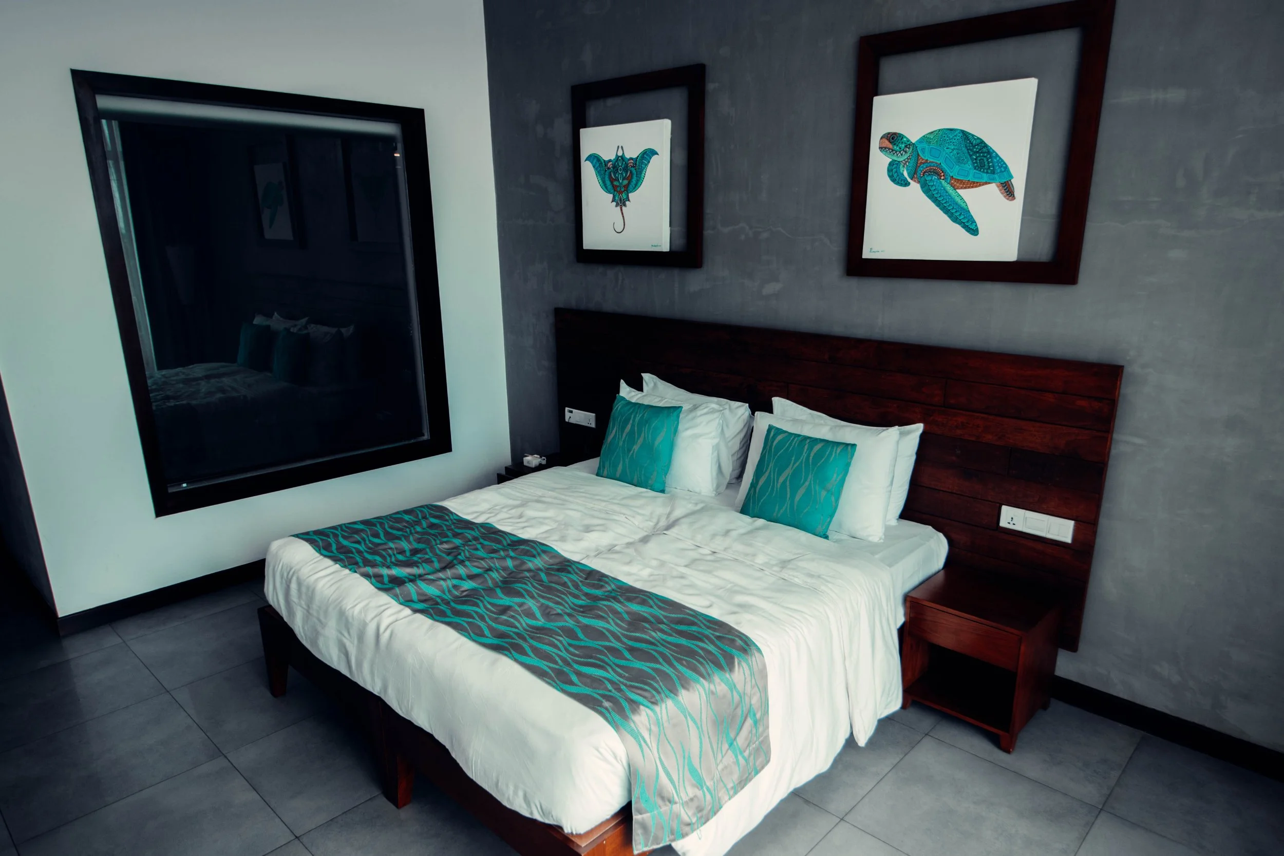 A bedroom with a large bed, two pillows with teal pillowcases, and a teal bed runner. There are two framed artworks of a turtle and a turtle with a plant above the bed. The room has a large window reflecting the bed and artwork.