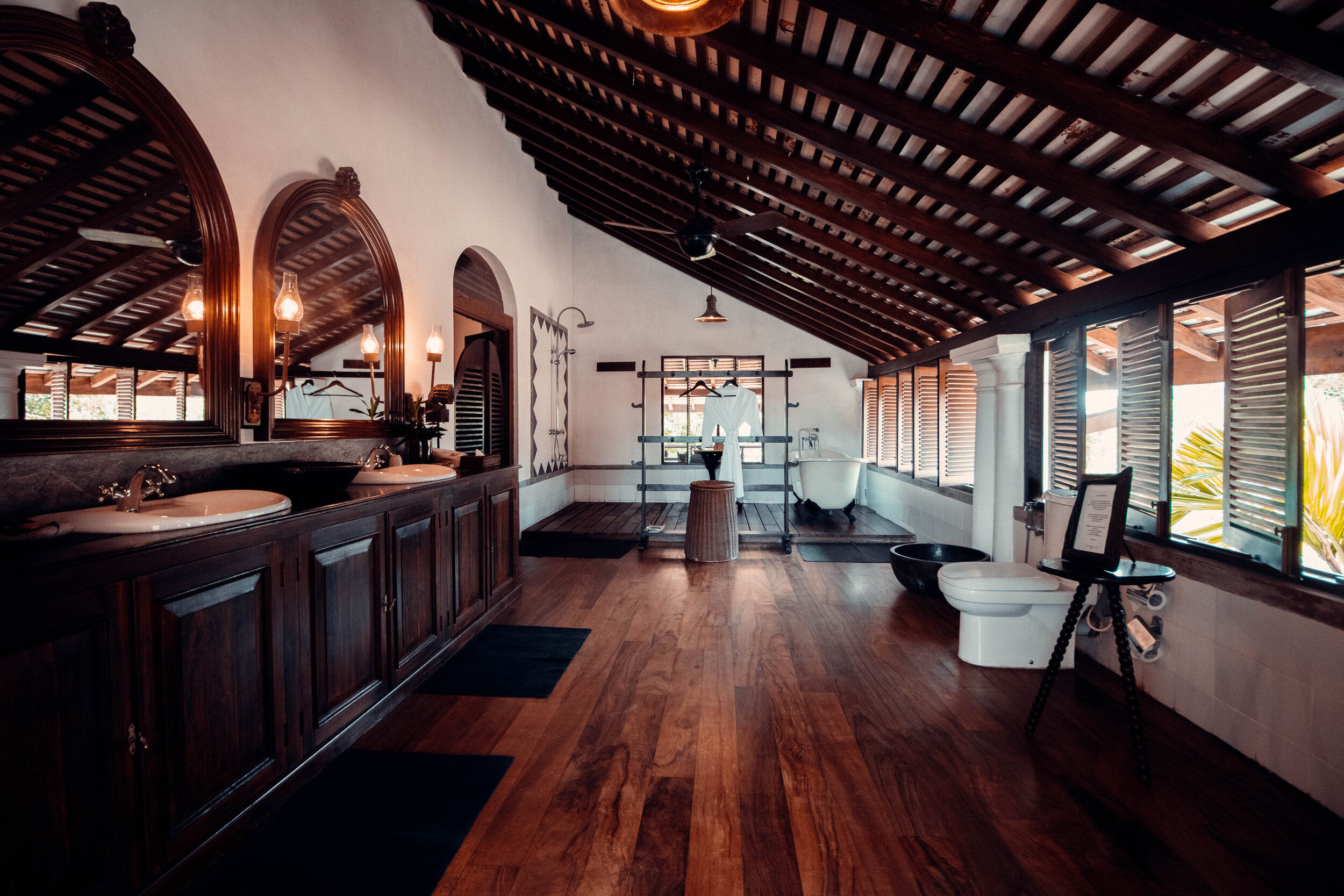 Interior of a rustic bathroom or spa with wooden floors, double sinks with large mirrors, vintage white bathtub, toilet, and windows with wooden shutters.