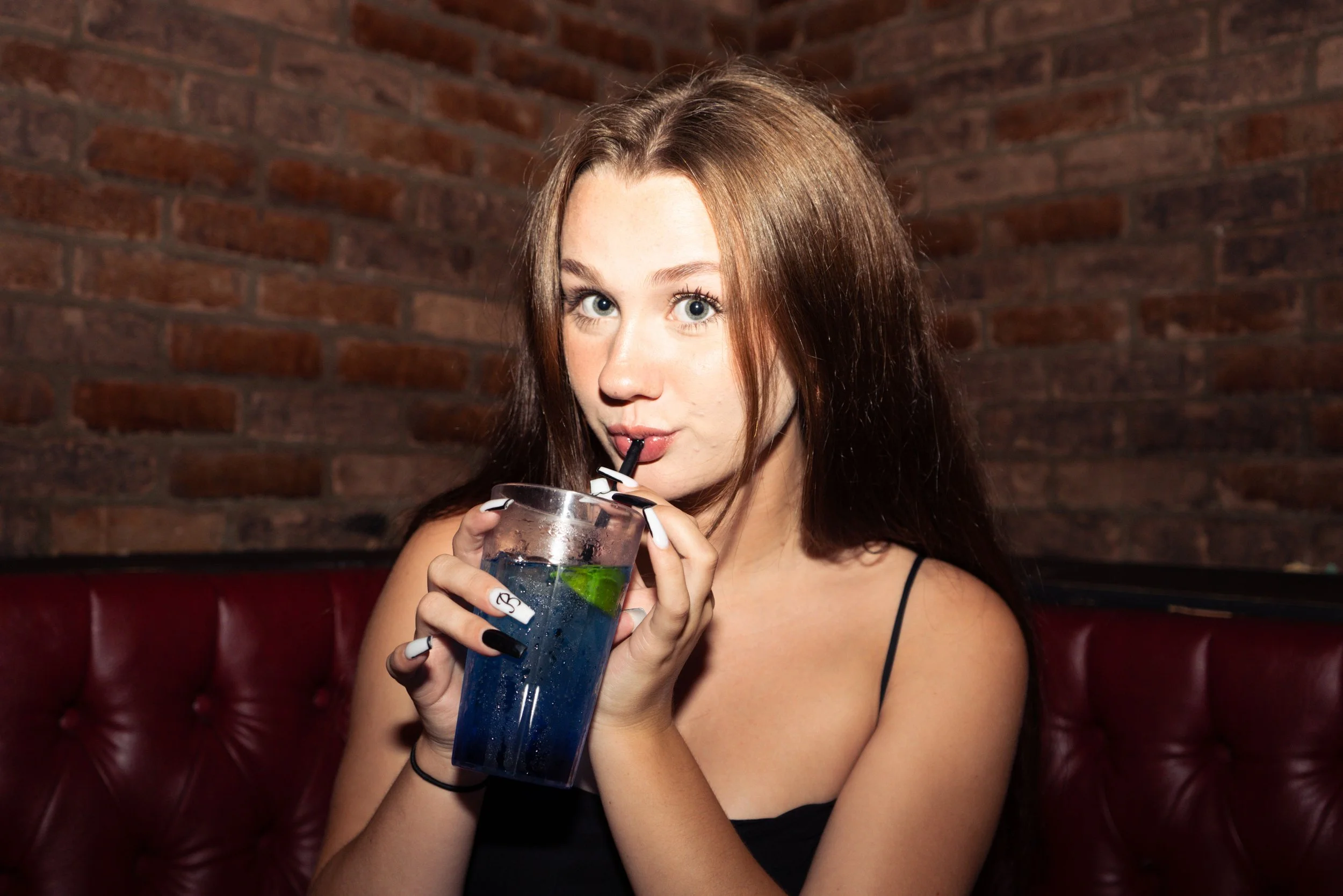 A young woman with long auburn hair and fair skin drinking a blue cocktail through a black straw in a glass with a lime wedge, sitting on a red leather booth with a brick wall background.