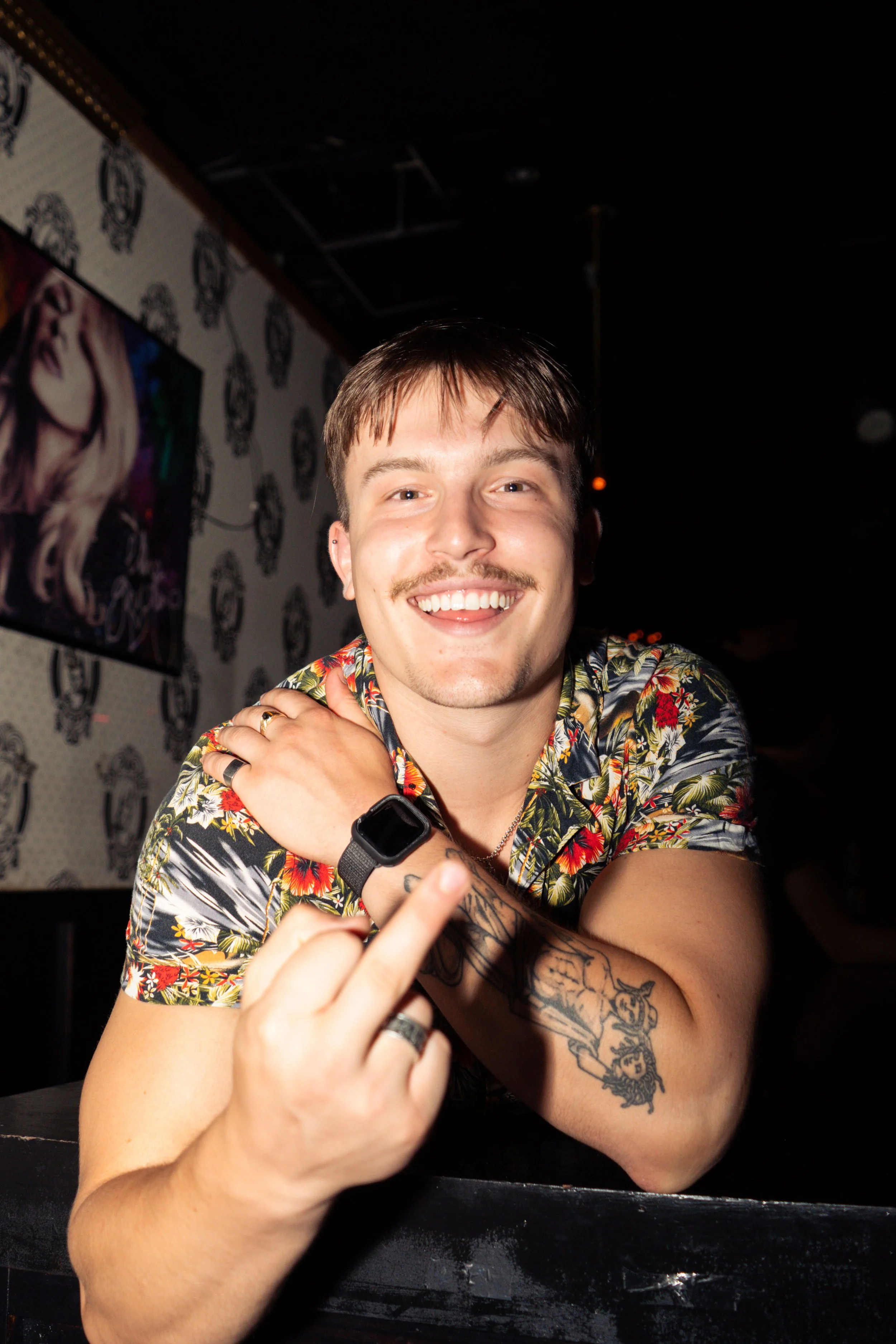 A smiling man with short brown hair and a mustache, wearing a floral shirt, showing middle finger at camera, sitting in a dimly lit bar or club.
