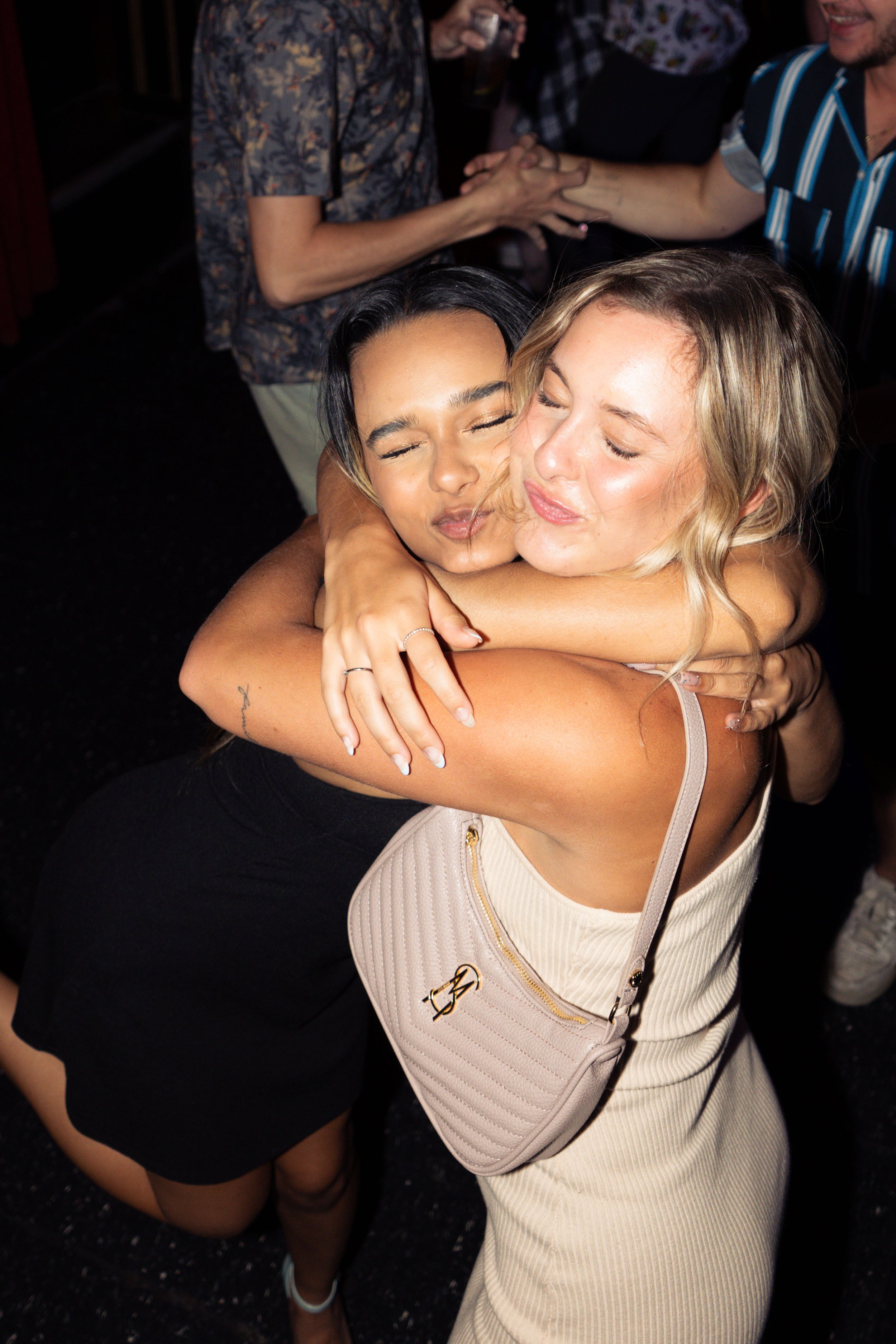 Two women hugging each other at a lively social gathering, smiling with eyes closed.