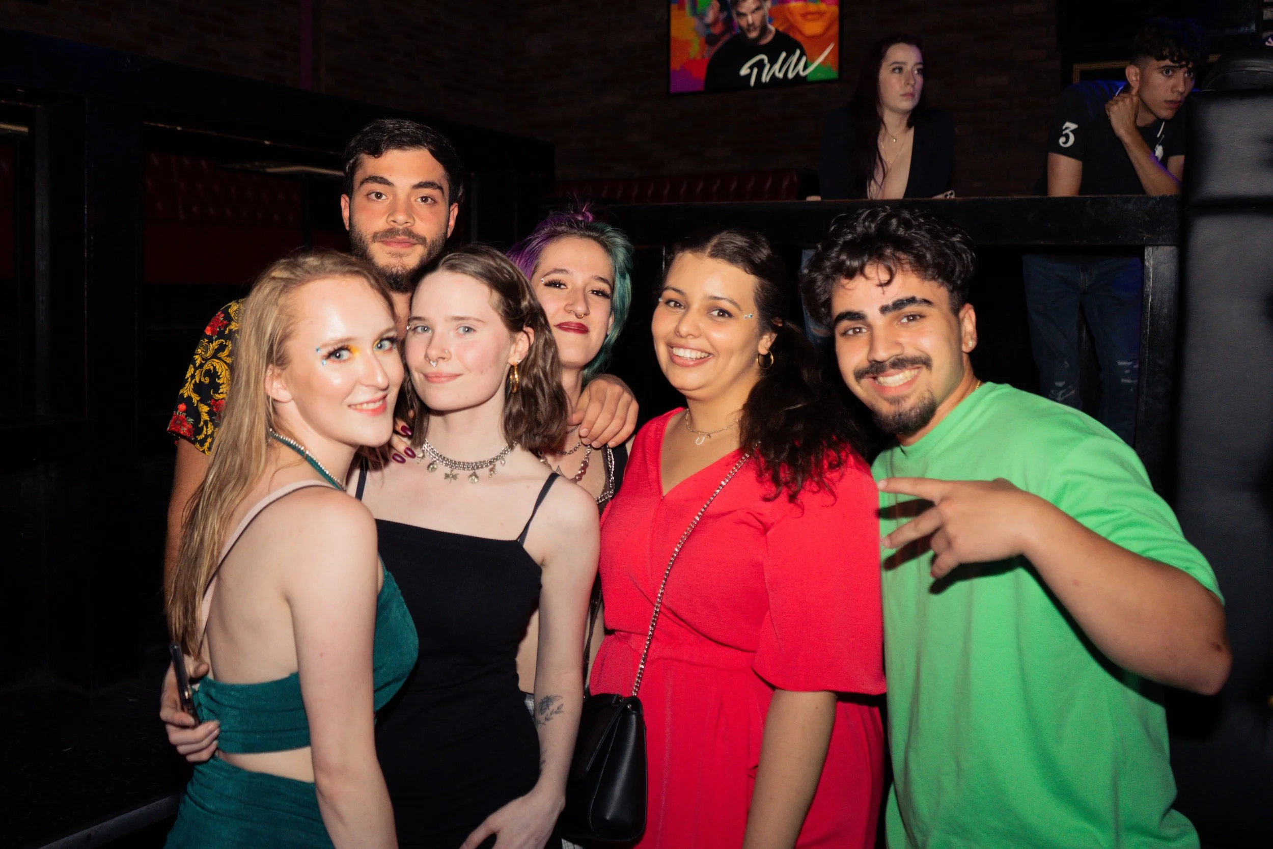 A group of seven young adults posing together at a social gathering or party, smiling at the camera, with a dark interior and some colorful artwork in the background.