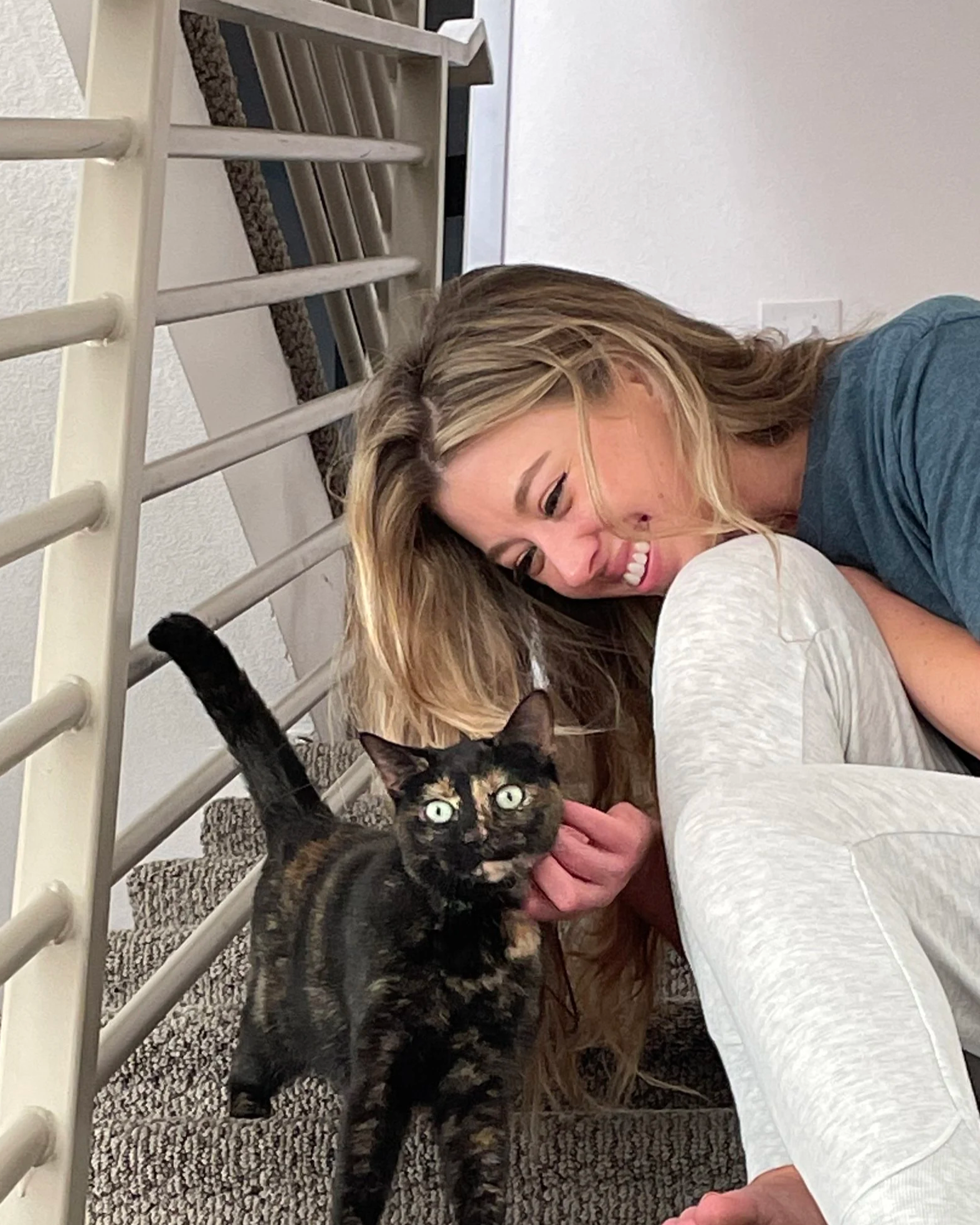 A woman with long blonde hair laughing while playing with a black and orange tortoiseshell cat on a gray carpeted staircase.