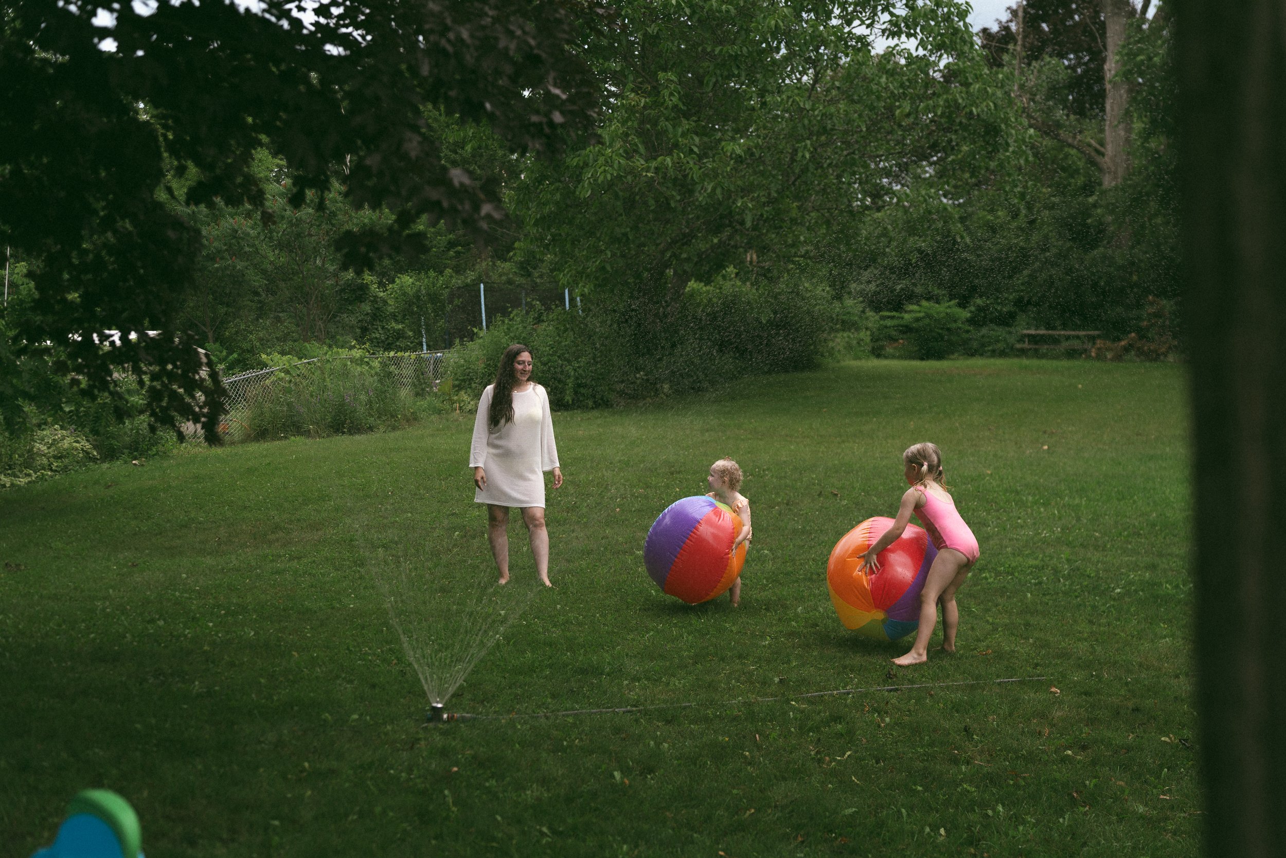 A woman and three children playing with large colorful beach balls in a backyard on a green lawn, trees and a chain-link fence in the background, water being sprayed from a sprinkler.