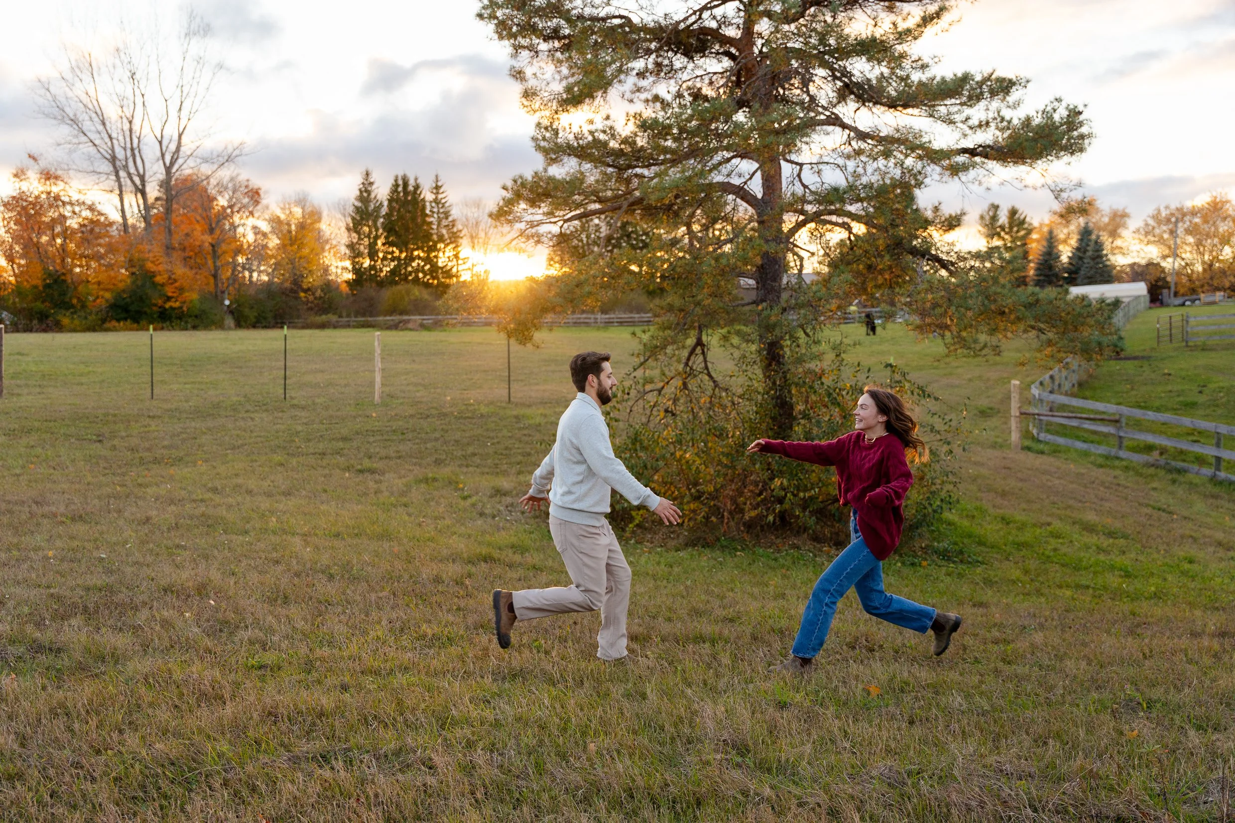 A young man and woman are playing and running outdoors in a grassy field during sunset. The woman is smiling and wearing a red sweater, and the man has a beard and is wearing a light-colored hoodie. There are trees with autumn foliage, a fence, and s