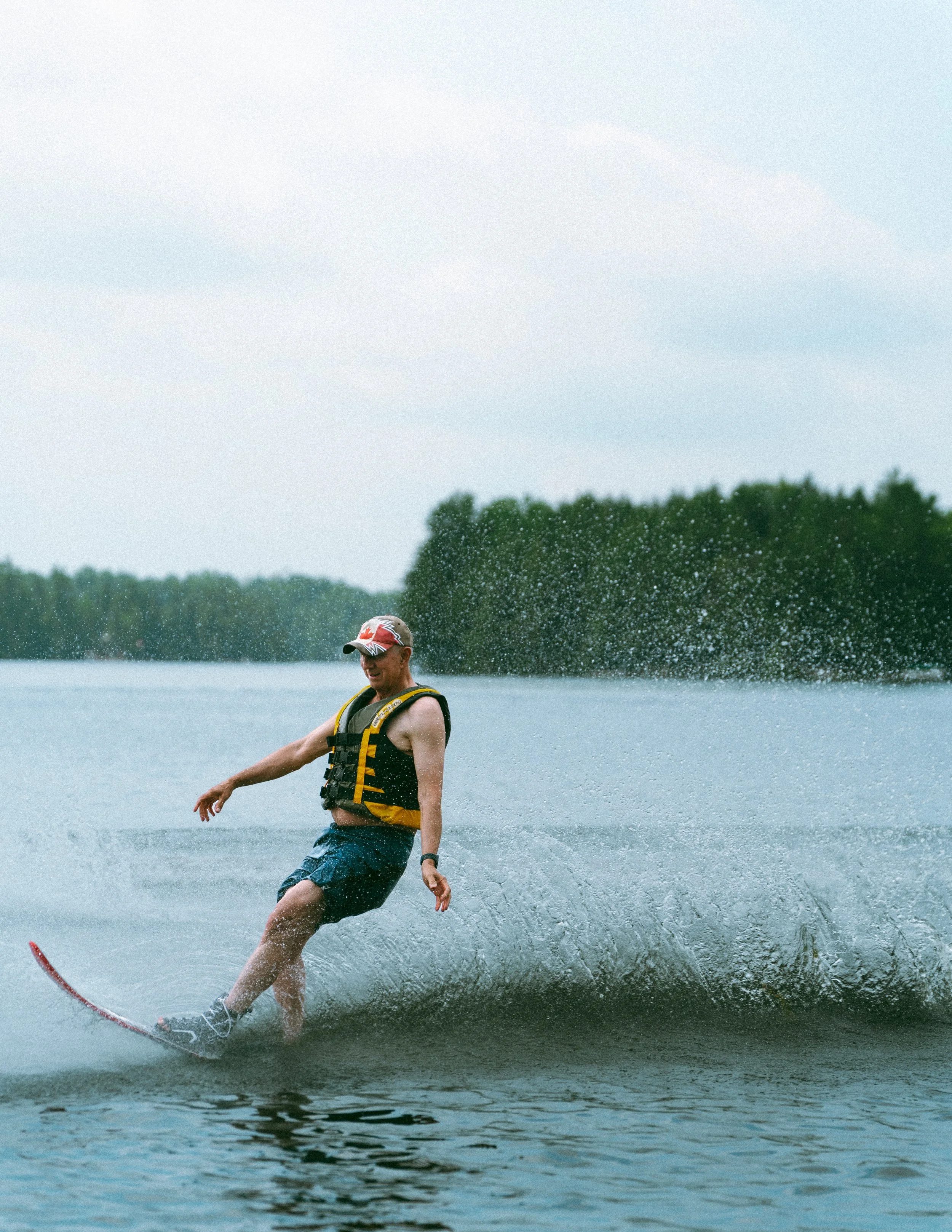 A man waterskiing on a lake during daytime, wearing a life jacket and a cap, with trees and cloudy sky in the background.