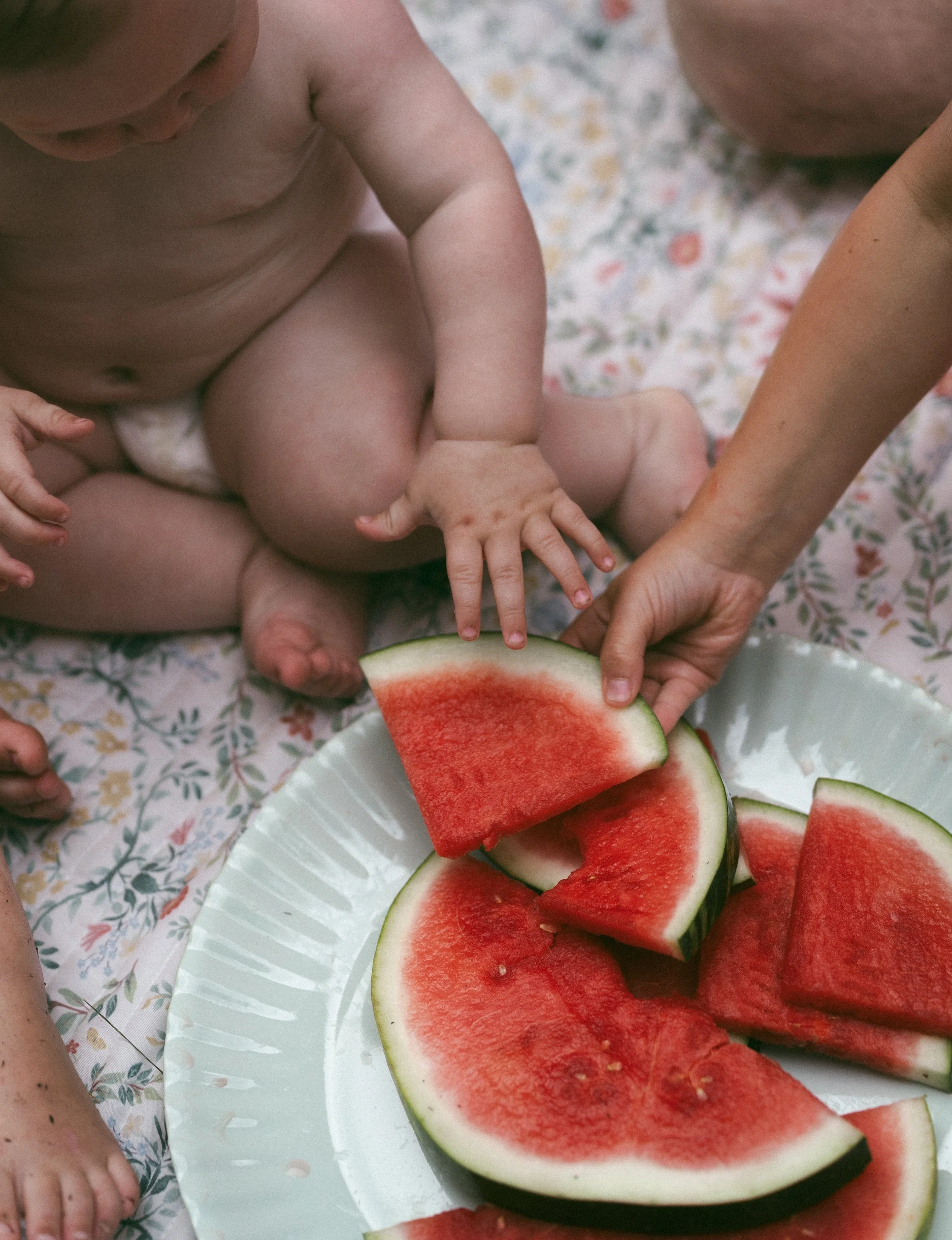 Close-up of a child's hand reaching for slices of watermelon on a white plate, with a patterned cloth underneath.