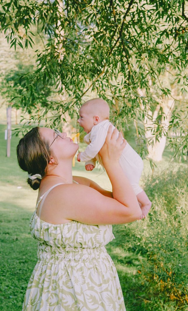 A woman holding a baby up in the air outdoors under green leafy trees, smiling and looking at each other.