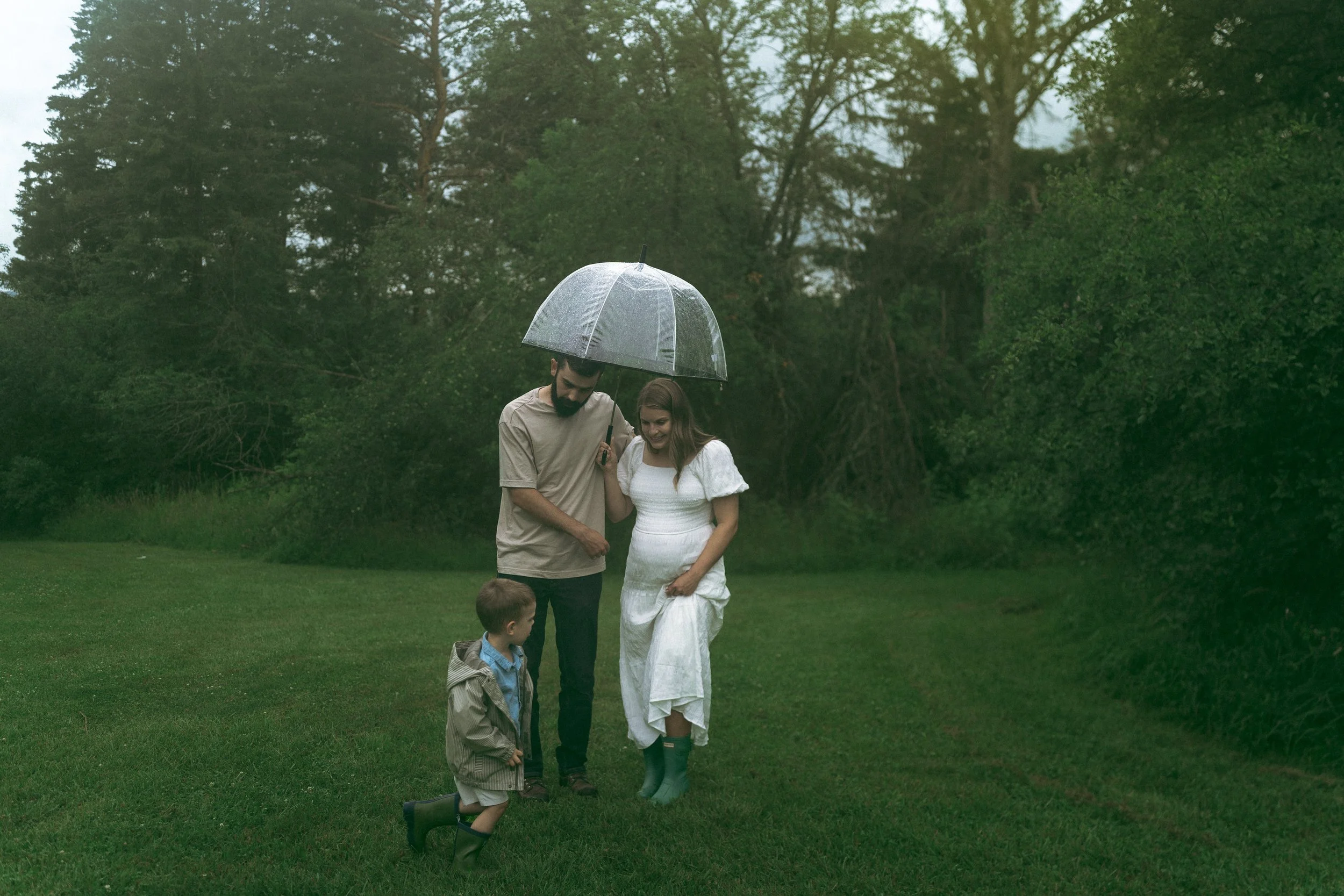 A man and a woman holding a transparent umbrella while standing on a grassy field on a rainy day, with two children nearby, all surrounded by green trees.
