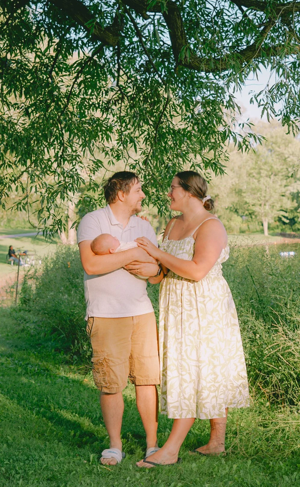 A baby being held by a man, with a woman standing opposite him in a park with green trees and grass, sharing a joyful moment.
