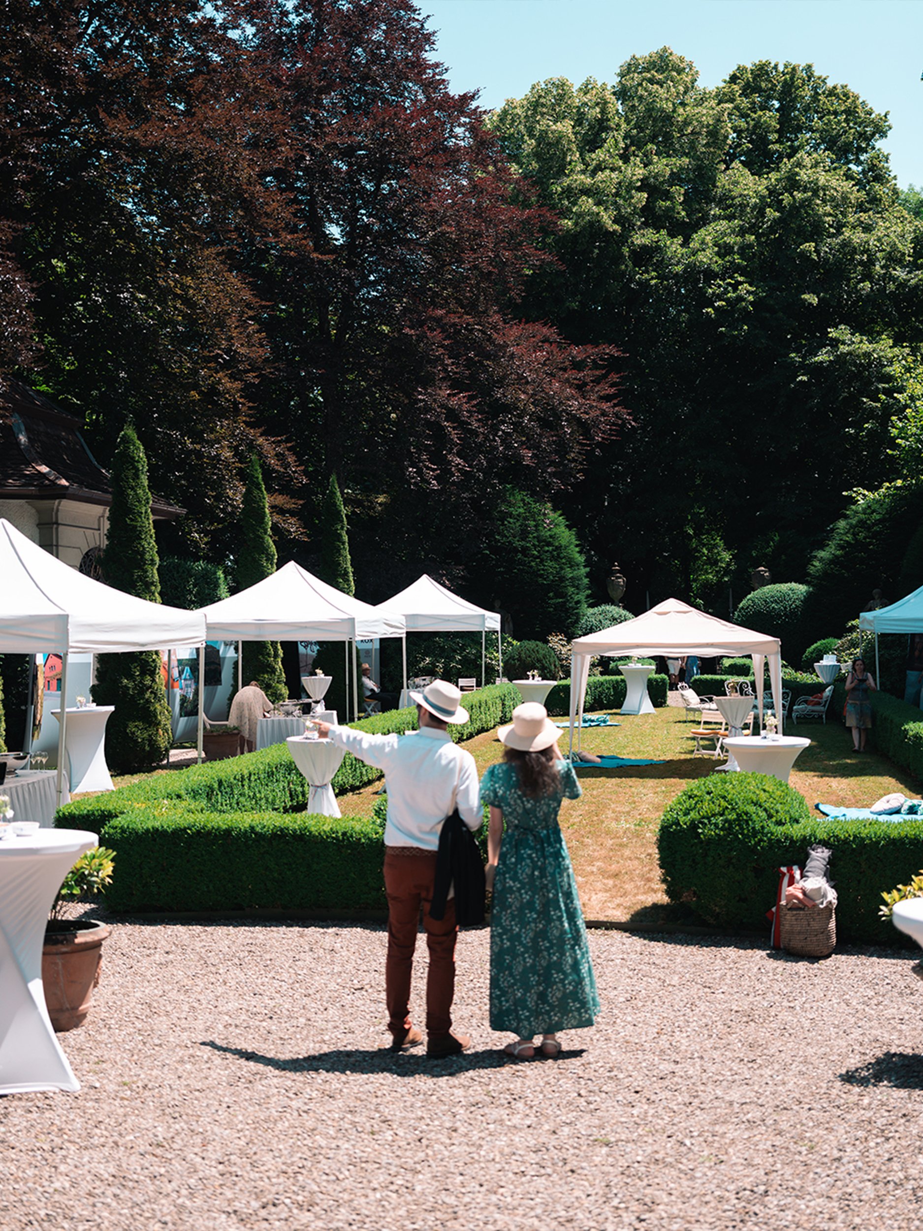 Sommerliches Gartenevent in einem Schlosspark mit weißen Pavillons und Stehtischen.