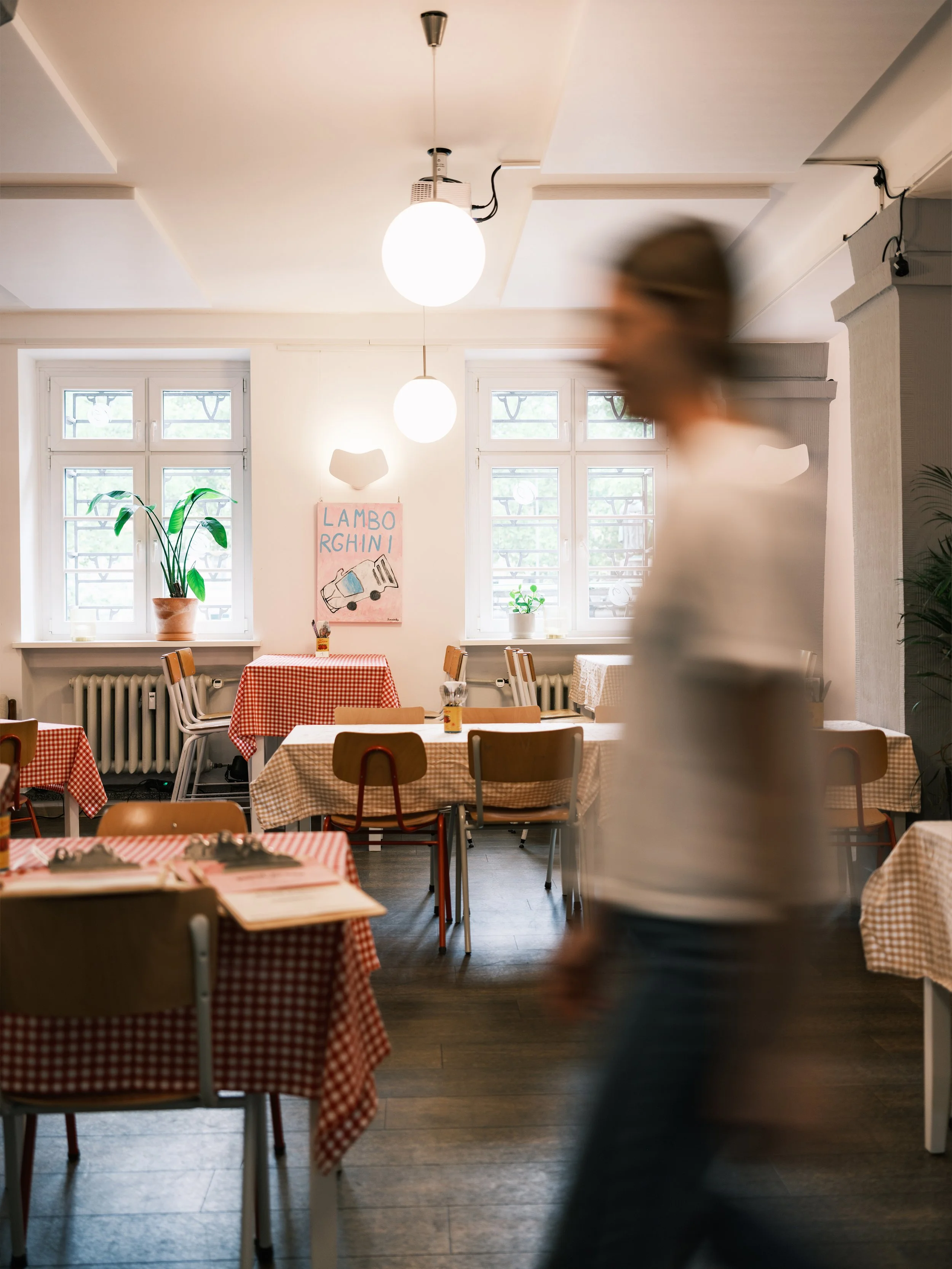 Ein heller Raum in einem Restaurant mit mehreren Tischen, die mit rot-weiß karierten Tischtüchern bedeckt sind, große Fenster, Pflanzen und eine Person im Vordergrund, die unscharf ist.