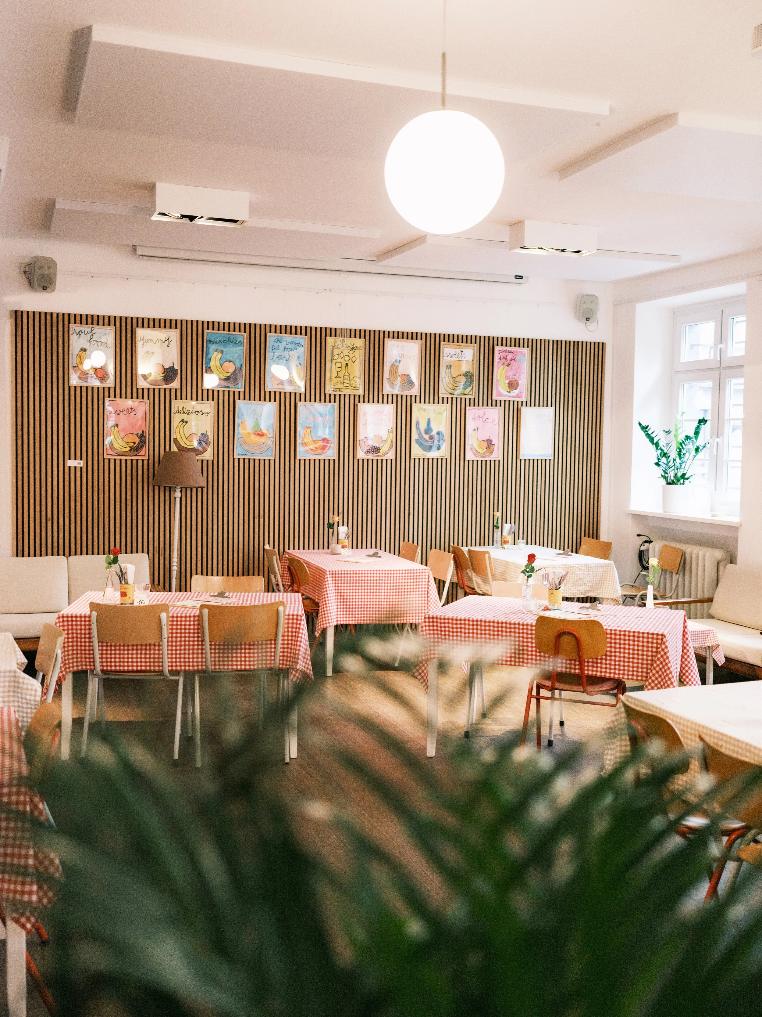 Ein heller, gemütlicher Essbereich in einem Café oder Restaurant mit mehreren Tischen, die mit rot-weiß karierten Tischtüchern bedeckt sind. Es gibt Zimmerpflanzen am Fenster und bunte, kindgerechte Wandbilder mit Früchten. Die Beleuchtung erfolgt durch Deckenleuchten und eine große, runde Deckenlampe.