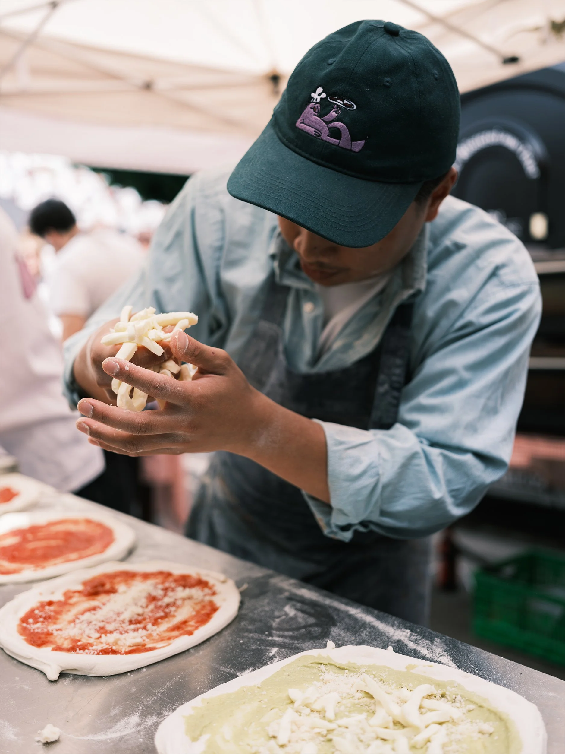 Pizzabäcker mit dunkler Cap belegt frische Pizzen mit Mozzarella, im Hintergrund ein Pizzaofen.