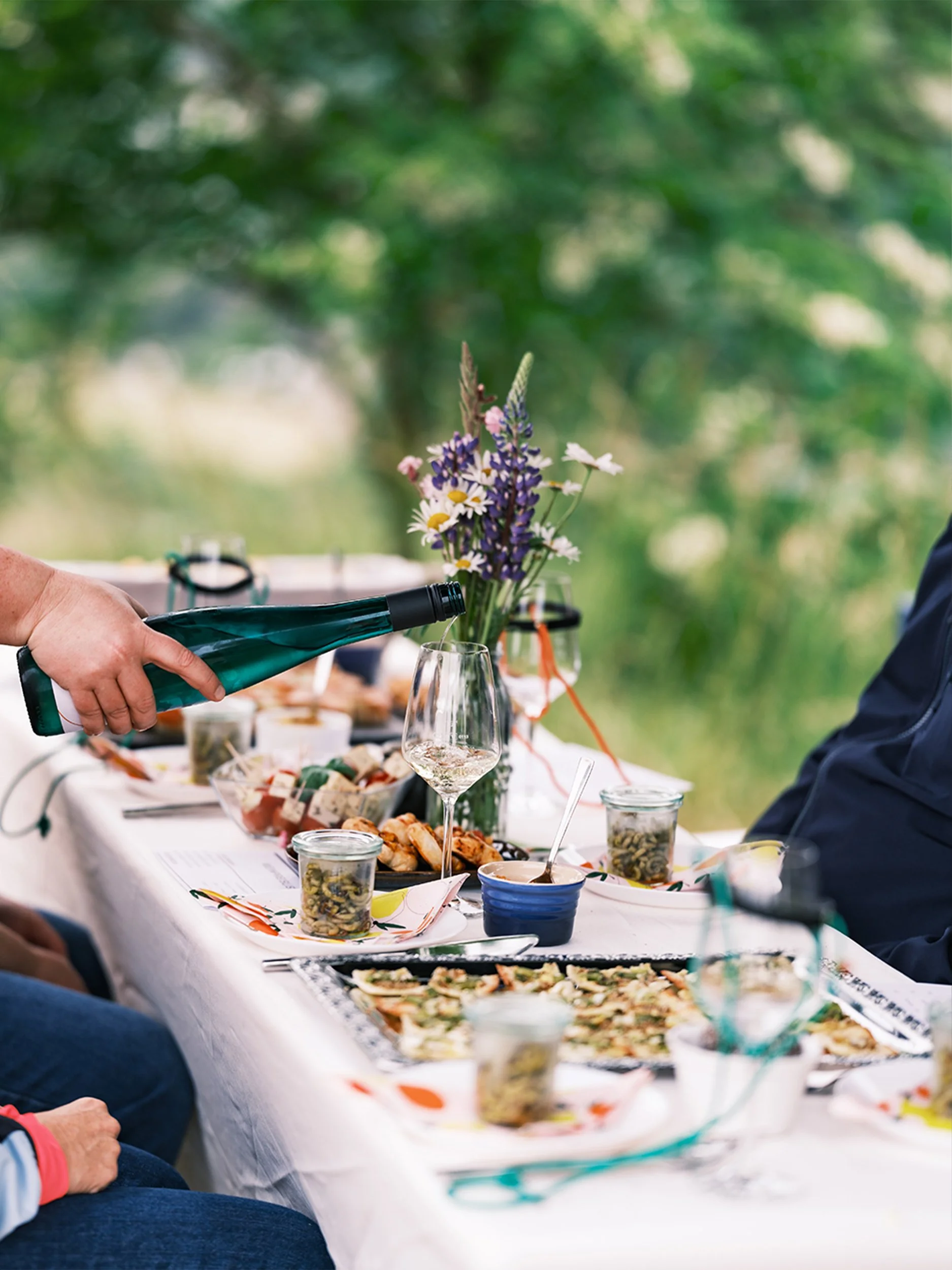 Hand schenkt Weißwein an einer sommerlich gedeckten Tafel im Grünen ein, dazu kleine Vorspeisen.