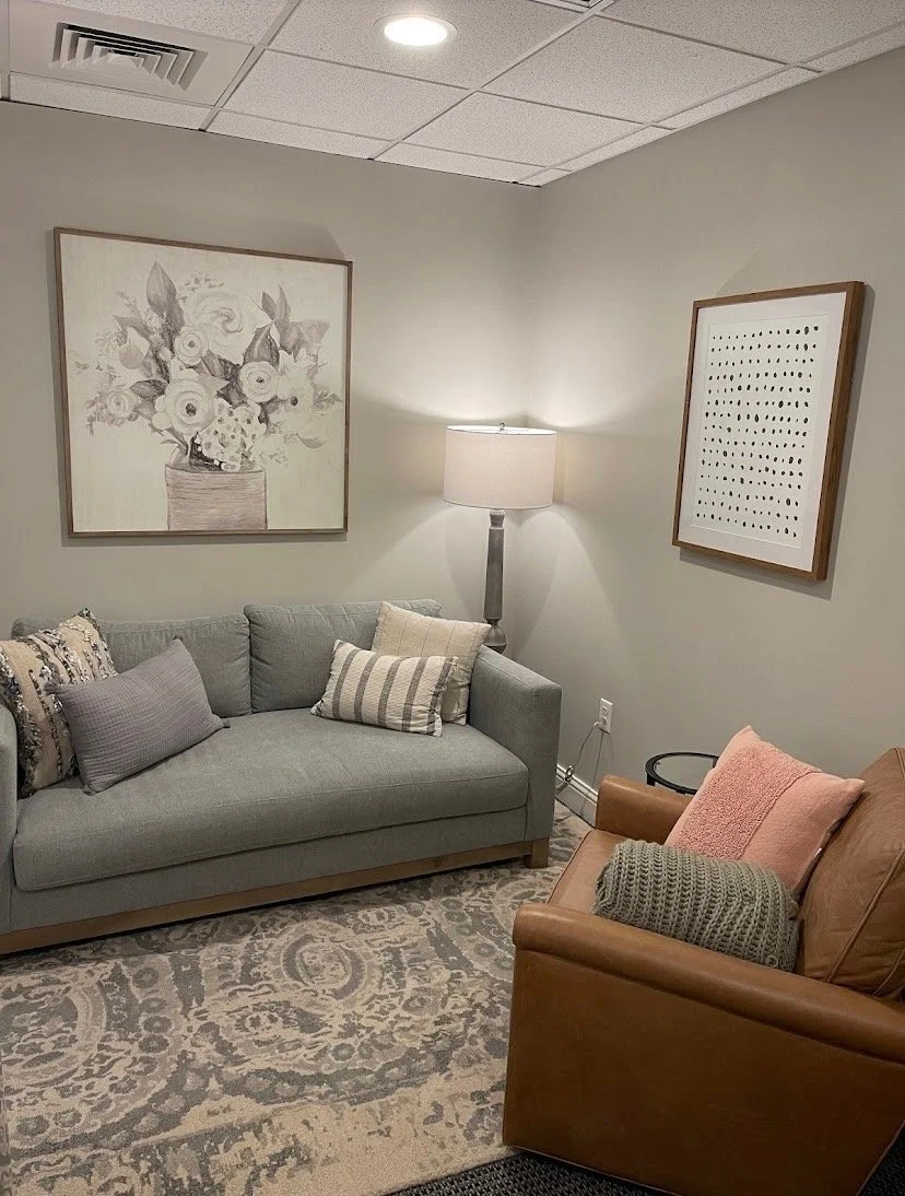 A cozy sitting room with beige walls, featuring a light gray sofa with multiple pillows, a brown leather armchair with pillows, a patterned area rug, a standing lamp, and framed artwork on the walls.