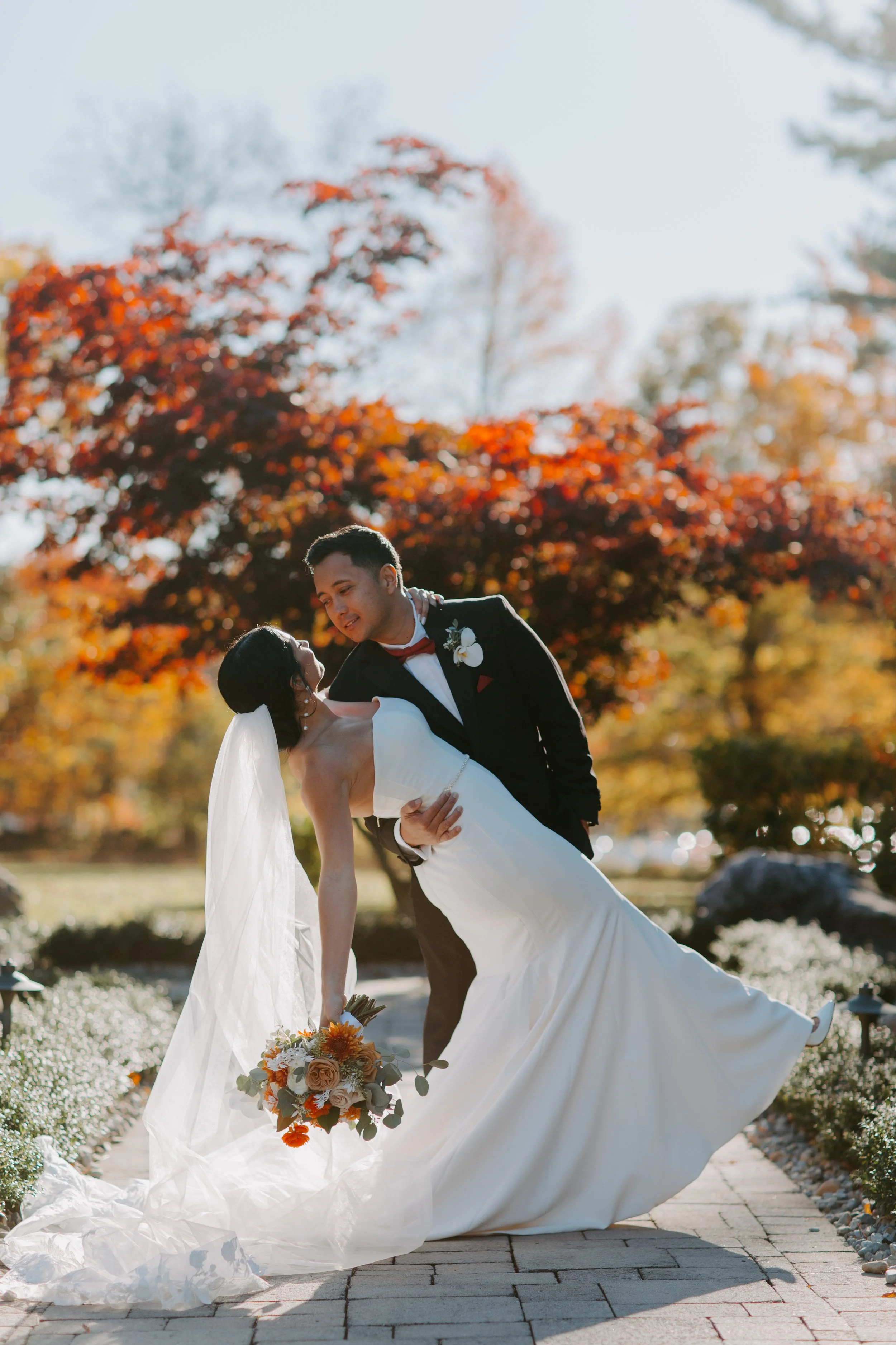 A bride and groom dancing outdoors in a park with autumn foliage.