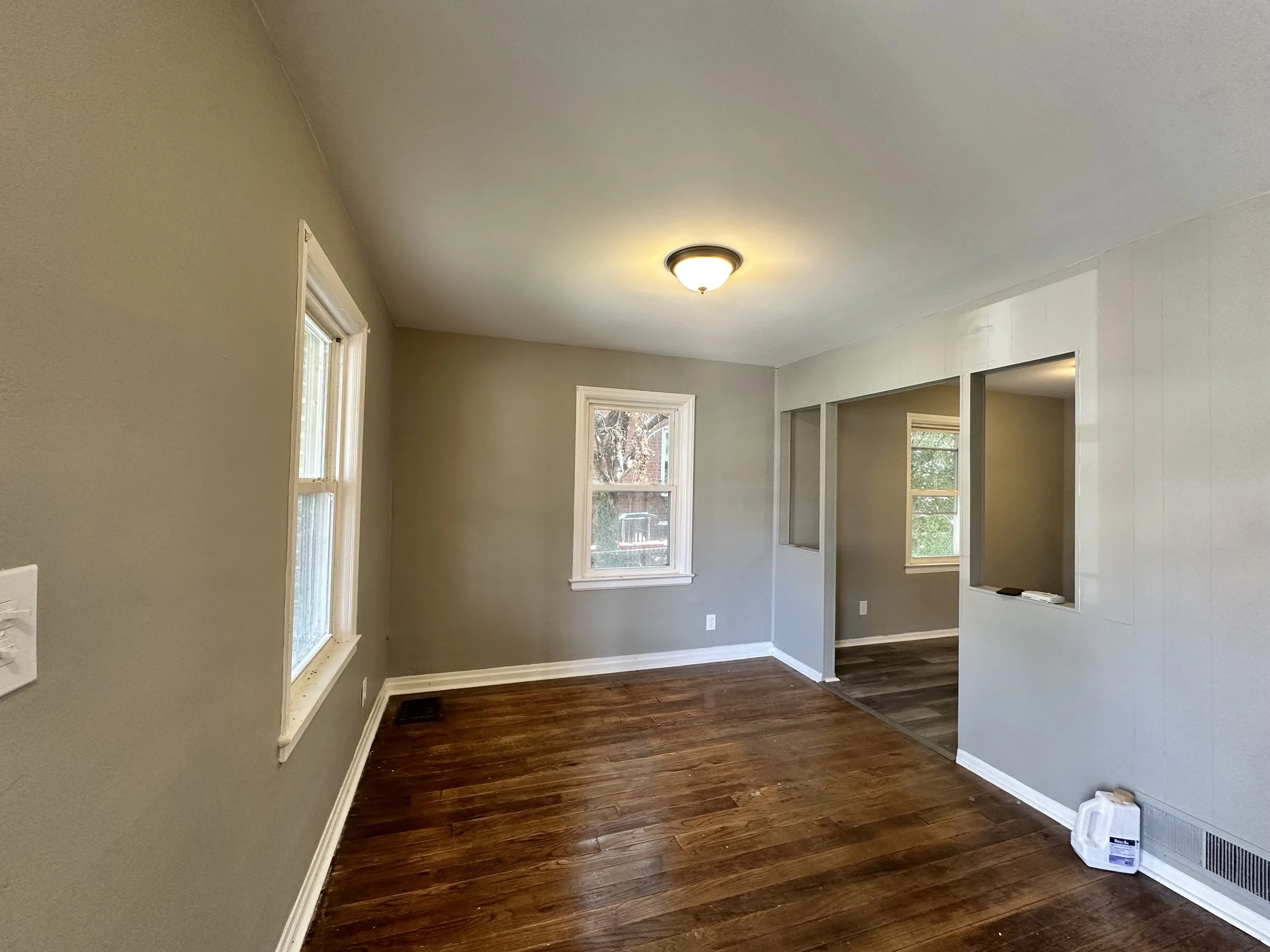 Empty living room with hardwood floors, light gray walls, three windows, and a ceiling light fixture.