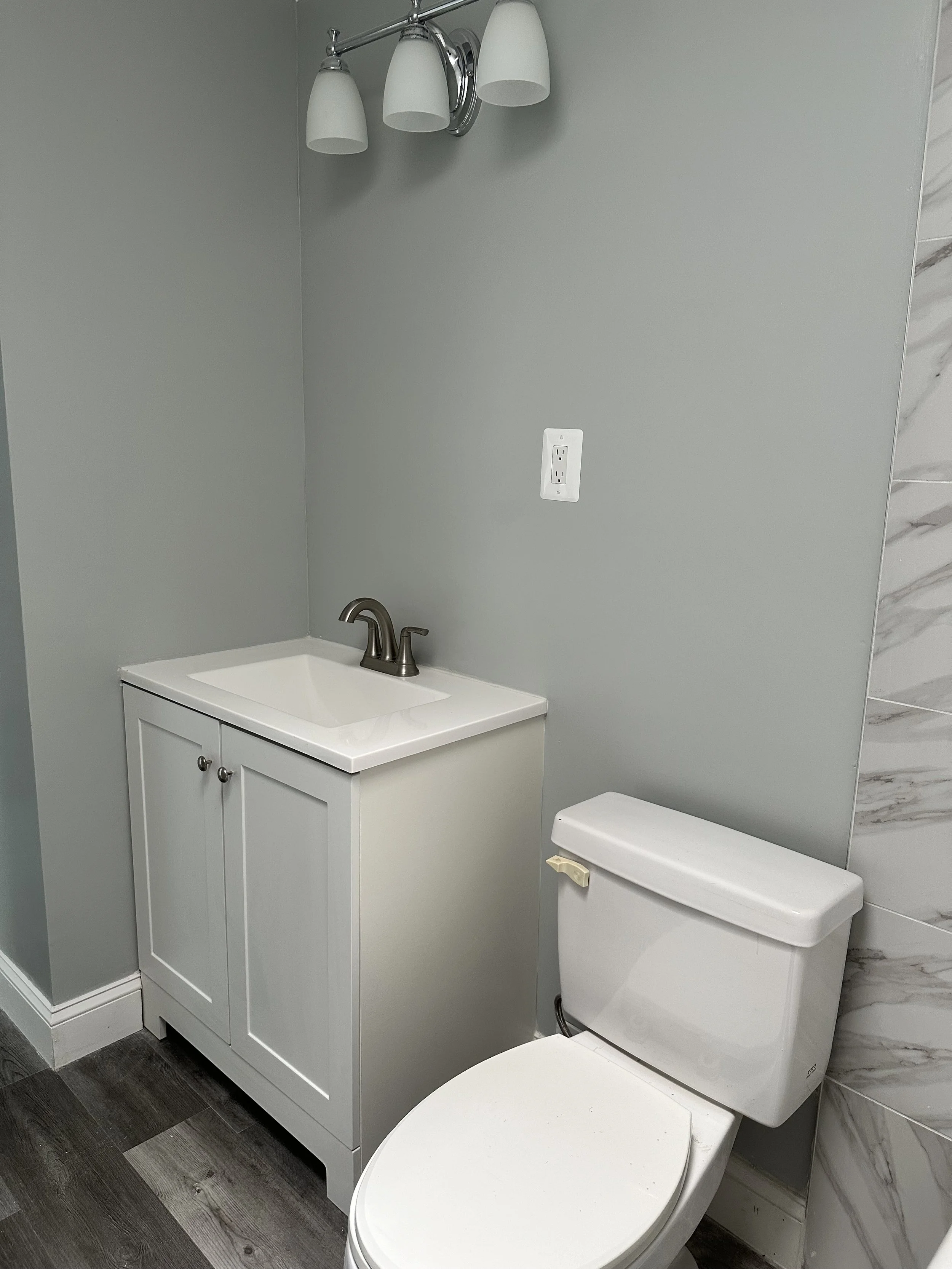 A small bathroom with a white sink cabinet, a silver faucet, a white toilet with a beige handle, a gray wall with an electrical outlet, a triple light fixture with white shades, and part of a tiled wall with a marble pattern.