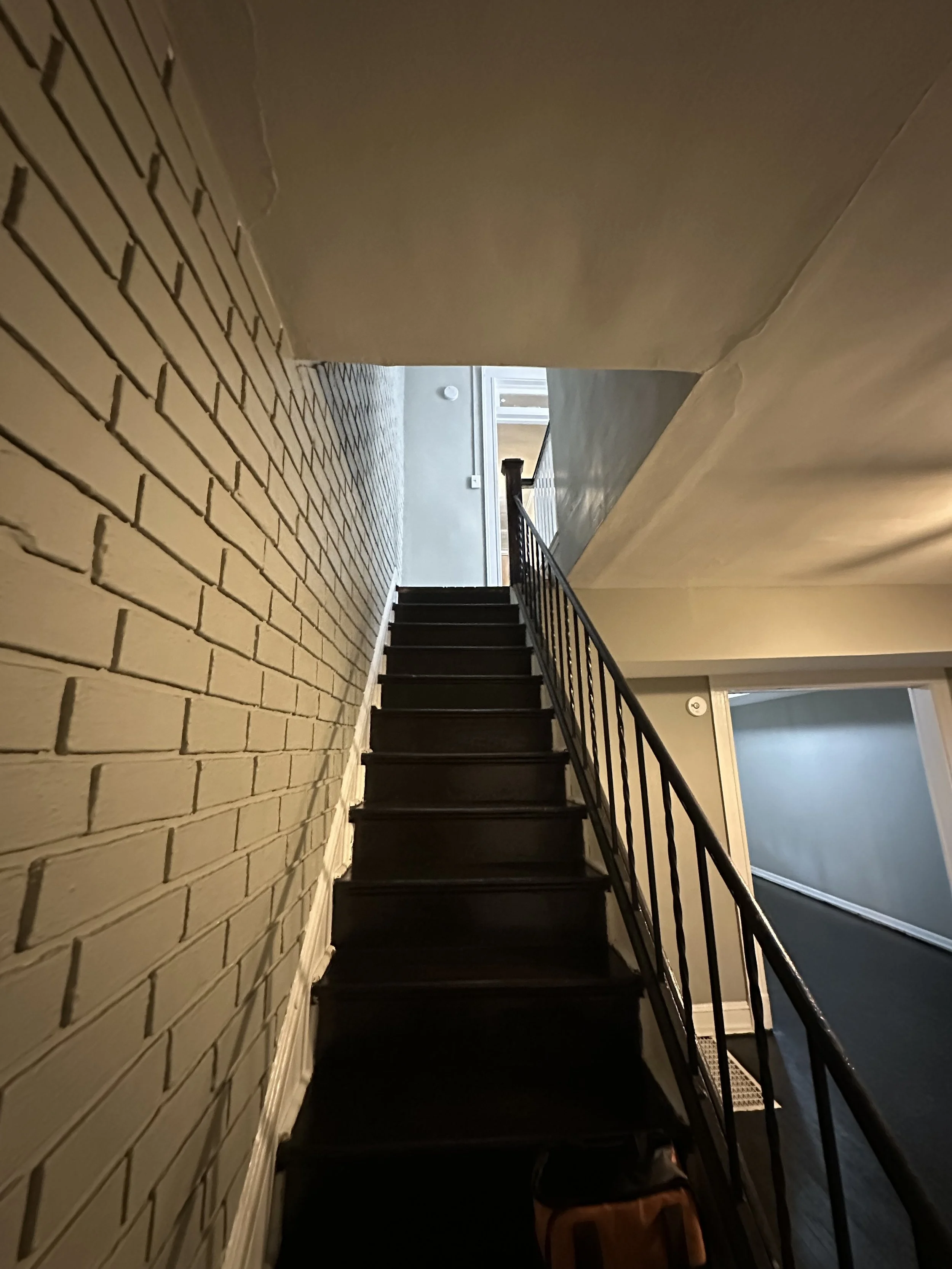 Interior staircase with black steps and a black metal railing, leading up to a second floor in a house with beige painted walls and a white brick wall on the left side.