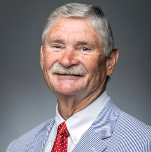 Close-up portrait of an elderly man with gray hair and a mustache, smiling, wearing a light blue suit, white shirt, and red tie, against a gray background.
