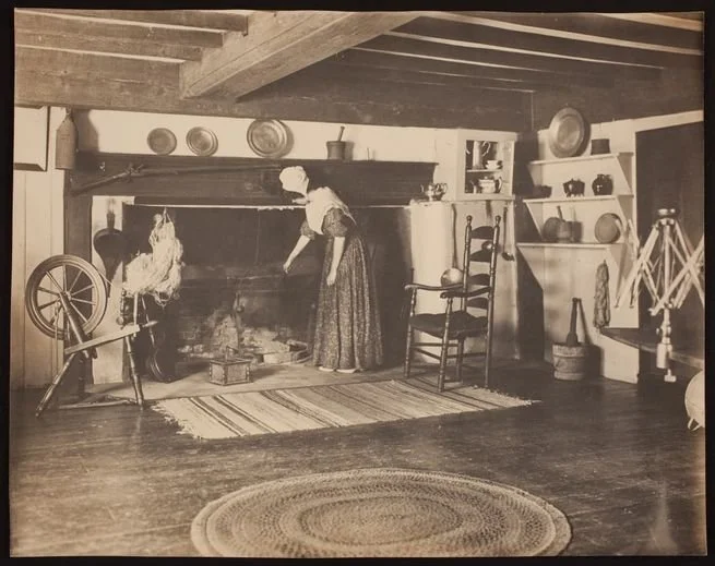 Black and white photograph of a woman in colonial-style dress, cooking at a fireplace.