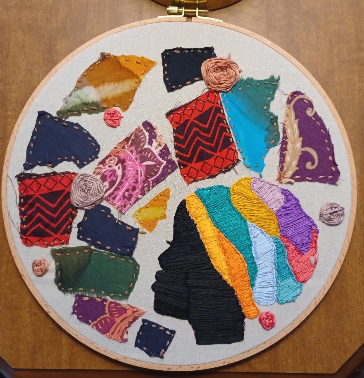 An embroidery piece, showing a silhouette of a woman in profile, wearing a colorful embroidered headscarf. She is surrounded by scraps of fabric stitched with gold thread and embroidered rosettes in shades of pink, rose-gold, and lavender.