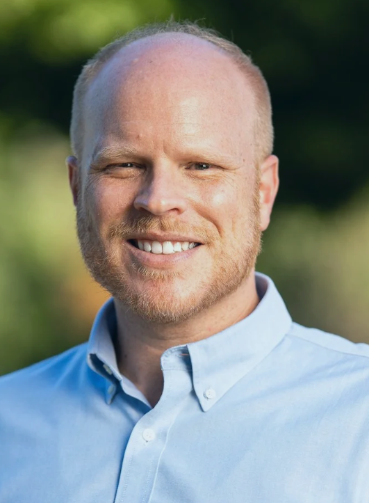 Photo depicting Zachary Dorholt from the shoulders up. He is a white male with a light red beard and red hair, smiling broadly. He is wearing a blue button-up shirt.