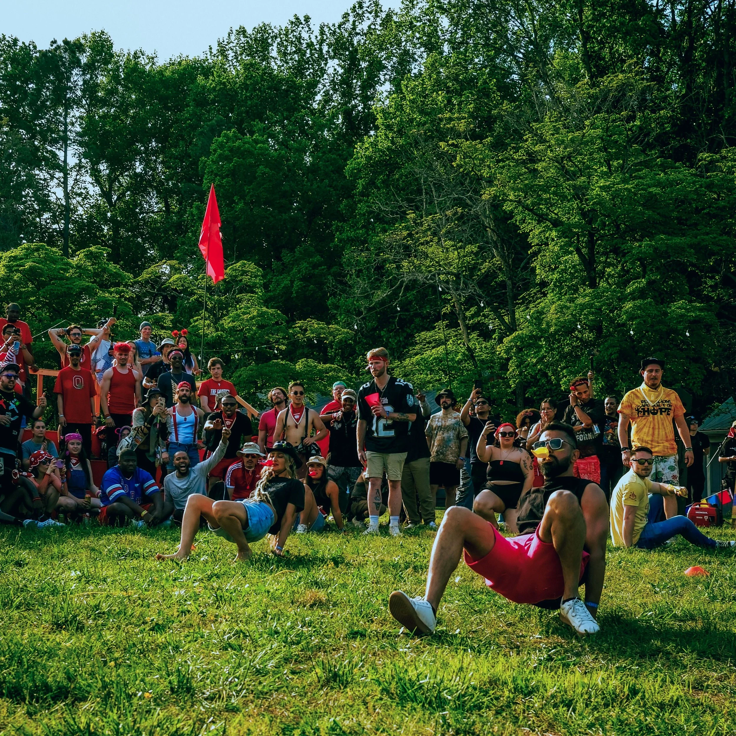 A diverse group of people gathered outdoors in a park, participating in an event, with many wearing red and patriotic clothing. One person in the foreground is doing a handstand on the grass, while others are standing and watching. The background features lush green trees under a clear sky.