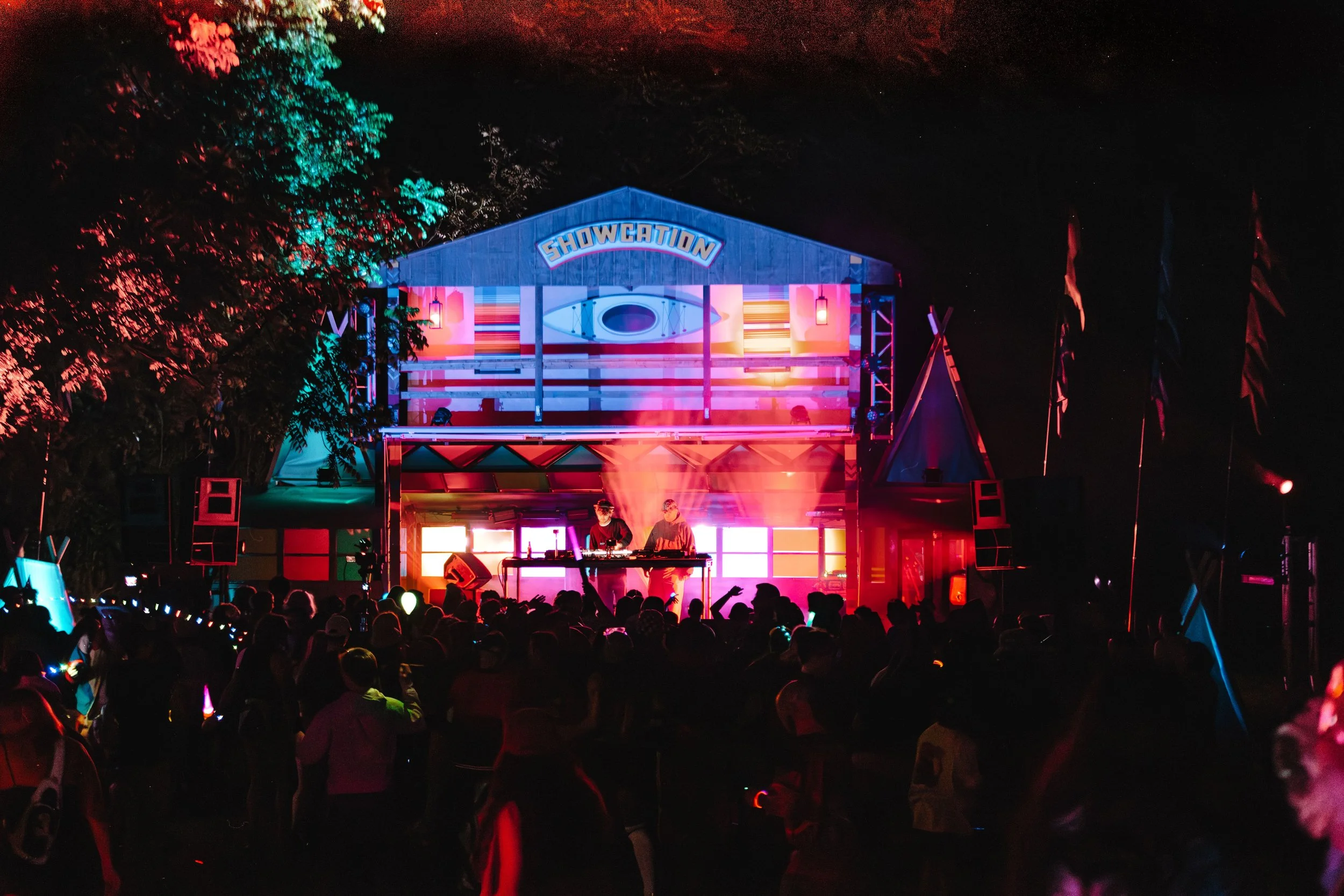 Nighttime outdoor concert with a stage labeled 'Showcation,' illuminated with colorful lights, featuring two DJs playing music, with a crowd of people dancing and enjoying the event.