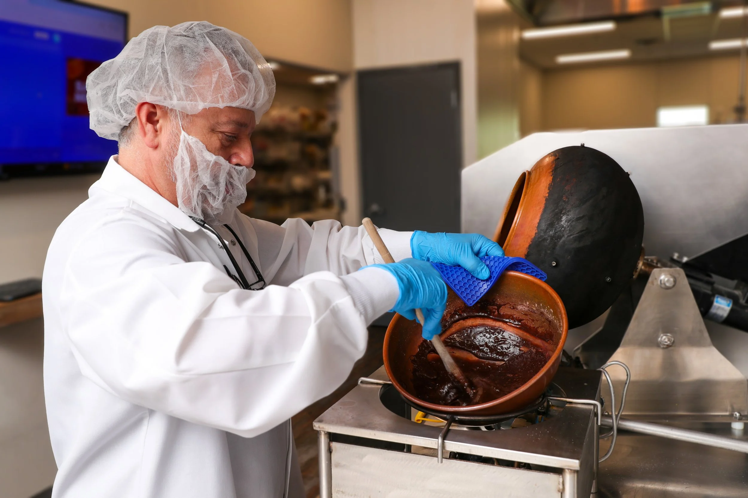 A man wearing a hairnet, face mask, white coat, and blue gloves stirring chocolate in a copper pot with a whisk in a commercial kitchen or chocolate laboratory.