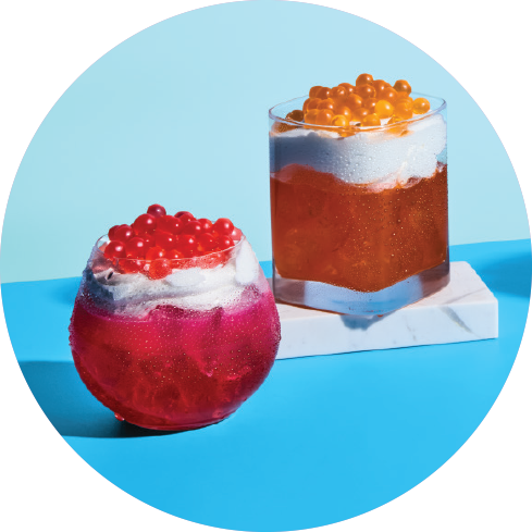 Two colorful bubble tea drinks with tapioca pearls, one in a round glass with pink and purple layers, and the other in a square glass with orange tapioca pearls and a white topping, set against a bright blue background.