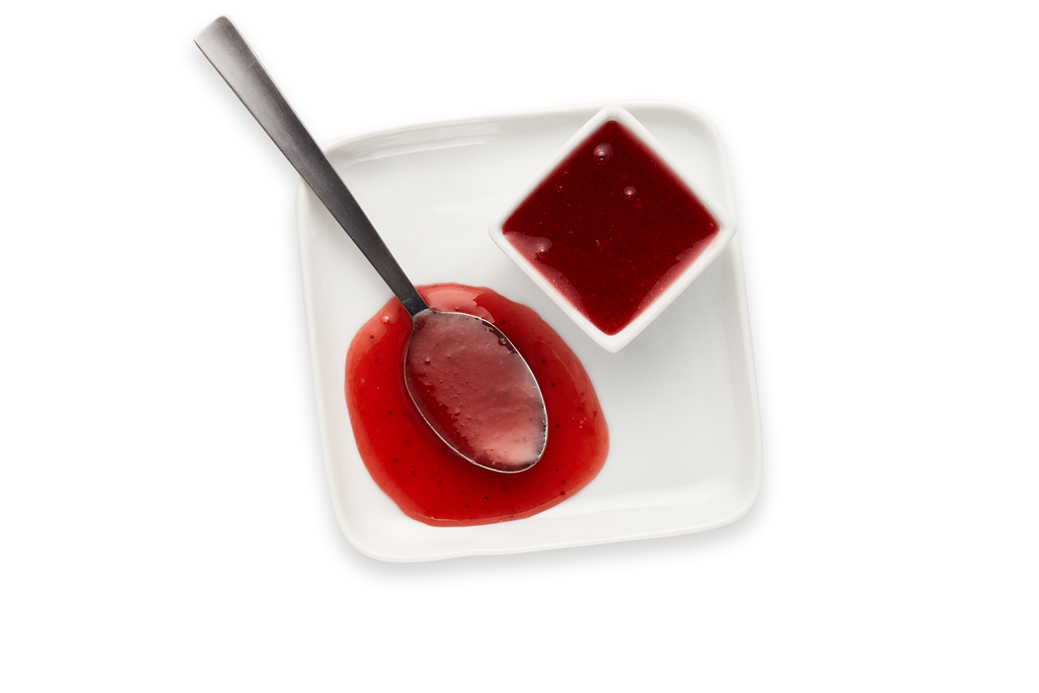 A white square dish contains a small white square bowl of red berry sauce and a metal spoon with some of the sauce, resting on a white surface.