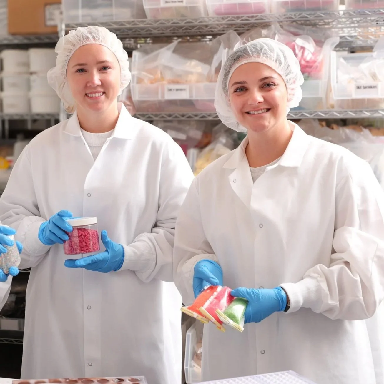 Two women in lab coats and hairnets smiling while holding colorful soaps in a storage room with shelves of supplies behind them.