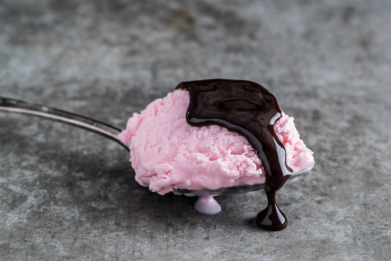A spoon holding a scoop of pink strawberry ice cream topped with chocolate syrup, on a gray textured surface.